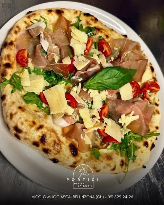 Pizza condita con prosciutto crudo, basilico, pomodorini e scaglie di formaggio su un piatto bianco.