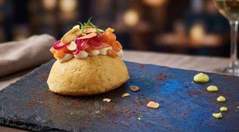 Taco gourmet su un piatto scuro, guarnito con verdure a dadini e salsa, con un pizzico di condimento blu sparso intorno.