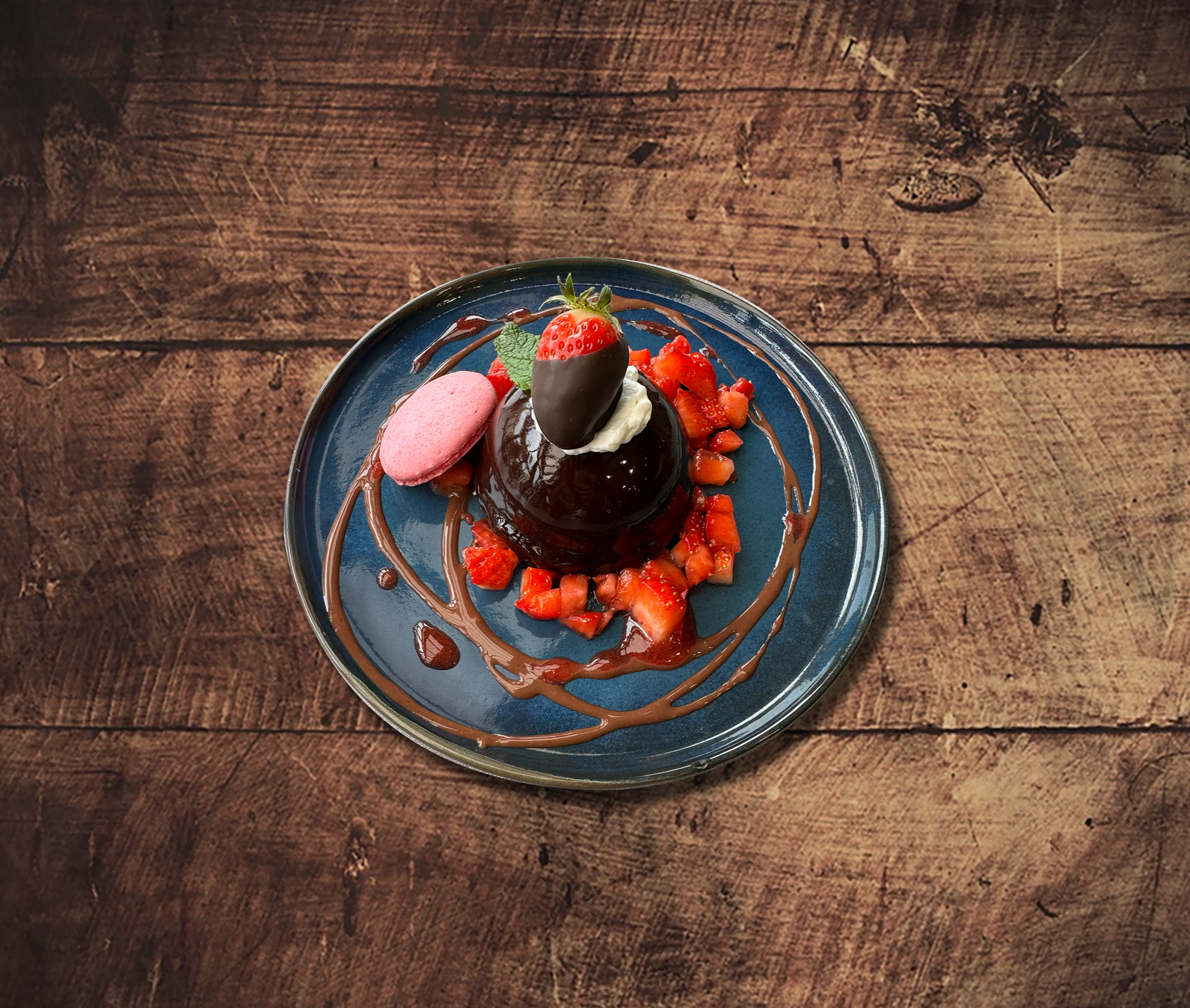 Dessert au chocolat avec fraises, macaron et sauce sur une assiette bleue, posée sur une surface en bois.