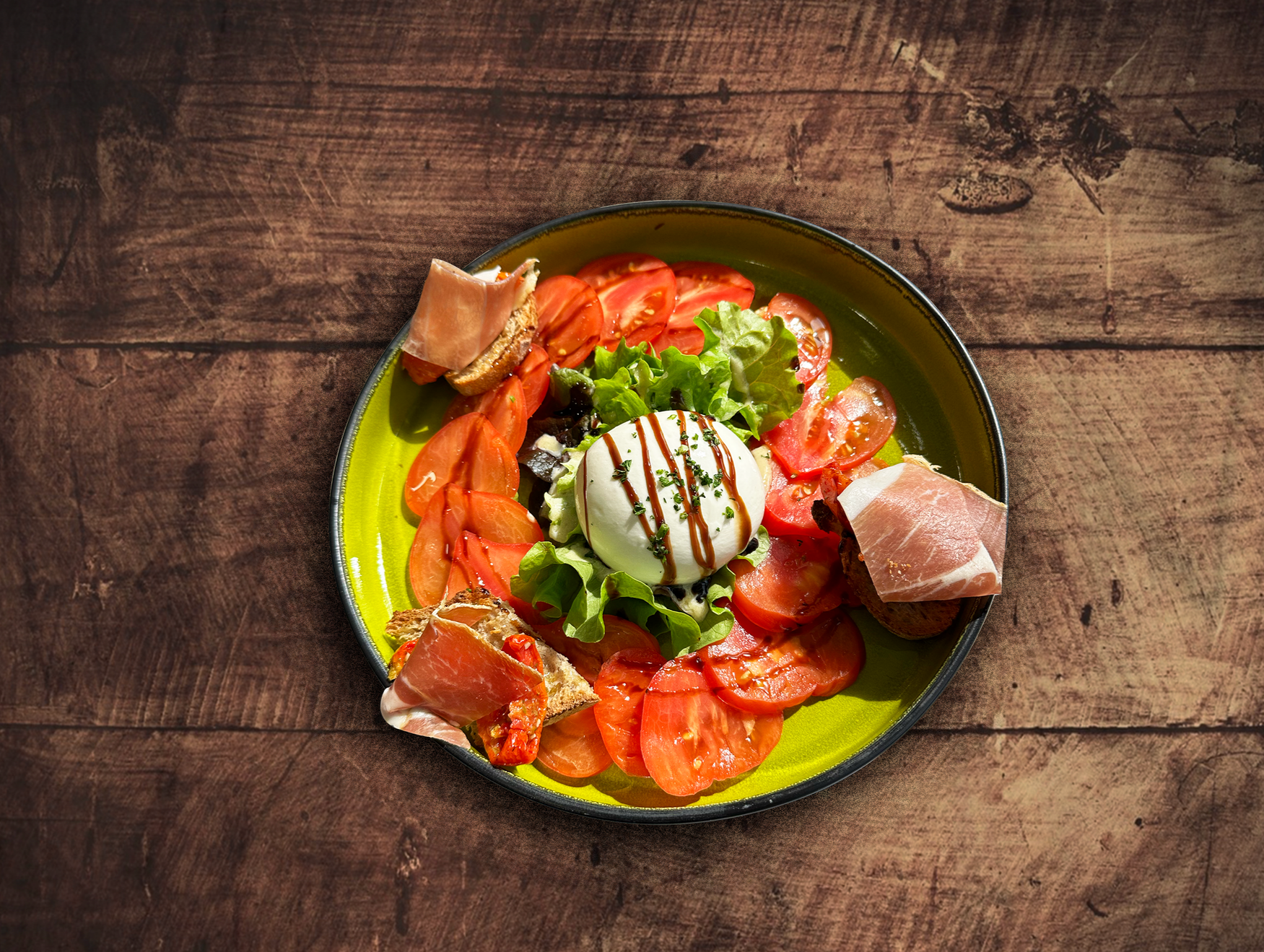 Assiette de tomates tranchées, de burrata, de laitue et de prosciutto sur une table en bois rustique.
