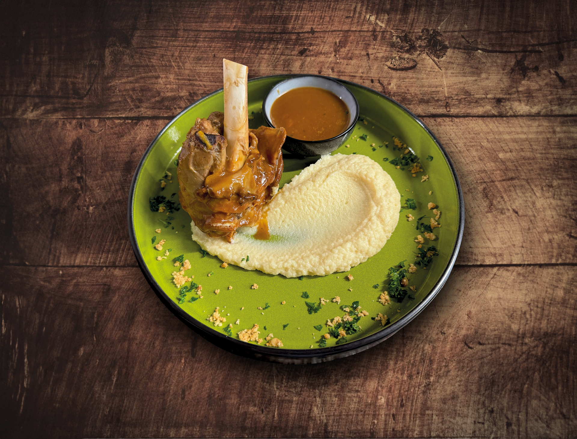 Jarret d'agneau avec purée de pommes de terre et sauce sur une assiette verte, table en bois.