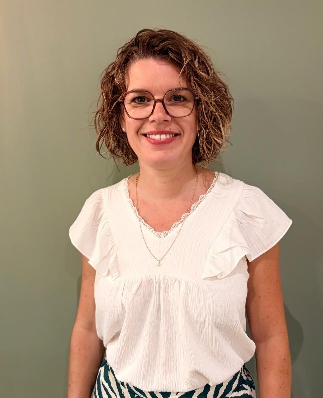 Femme souriante portant des lunettes et un chemisier blanc devant un mur vert uni.