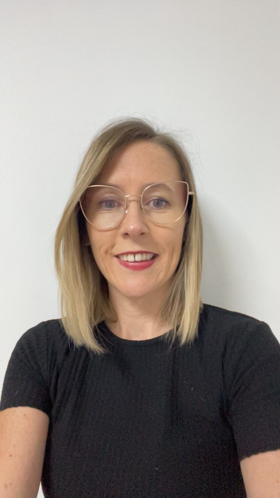 Portrait d'une femme souriante aux cheveux blonds et portant des lunettes, sur un fond clair uni.