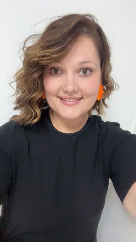 Personne souriante en chemise noire, cheveux ondulés et boucles d'oreilles orange, sur fond clair