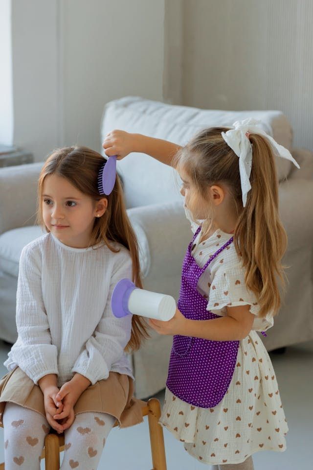 Deux enfants dans un salon ; l'un brosse les cheveux de l'autre avec un sèche-cheveux jouet.