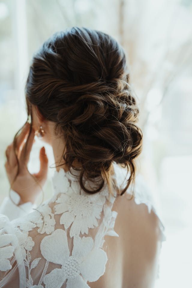 Femme coiffée d'un chignon élégant et vêtue d'une robe en dentelle, ajustant sa coiffure près d'une fenêtre lumineuse.