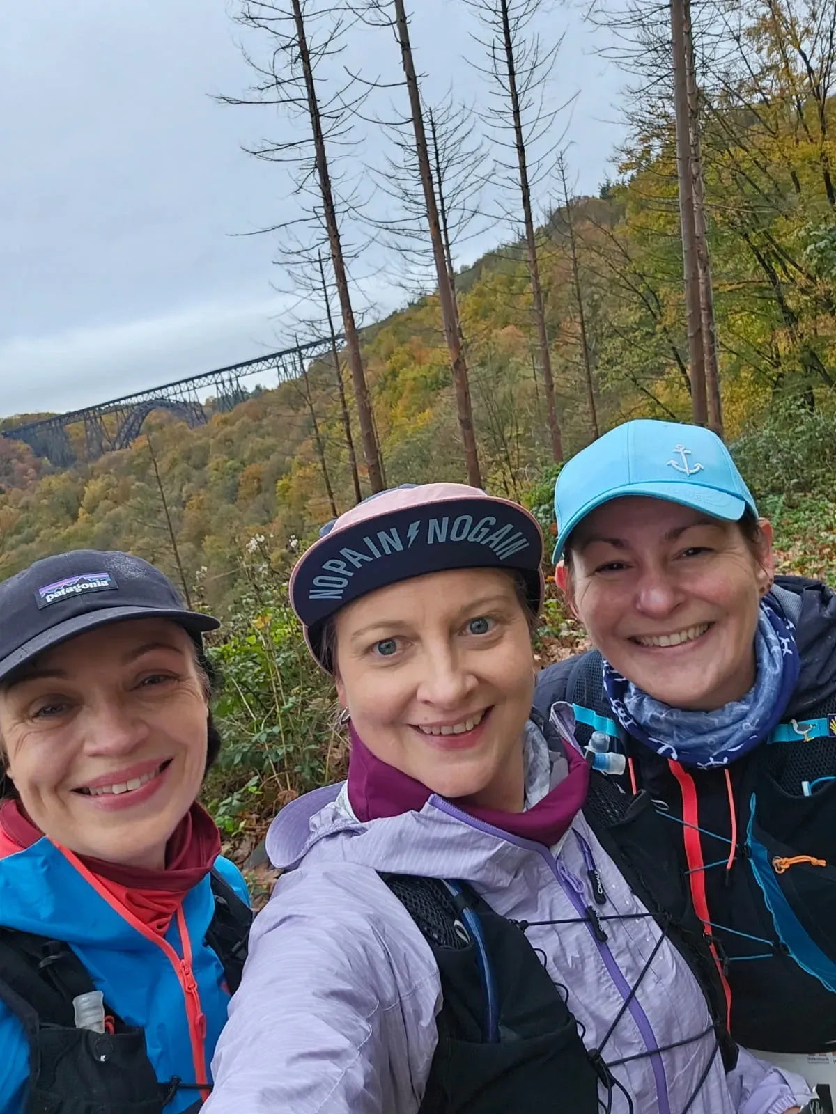 Drei lächelnde Frauen machen ein Selfie im Freien an einem Hang. Herbstlaub, bewölkter Himmel.