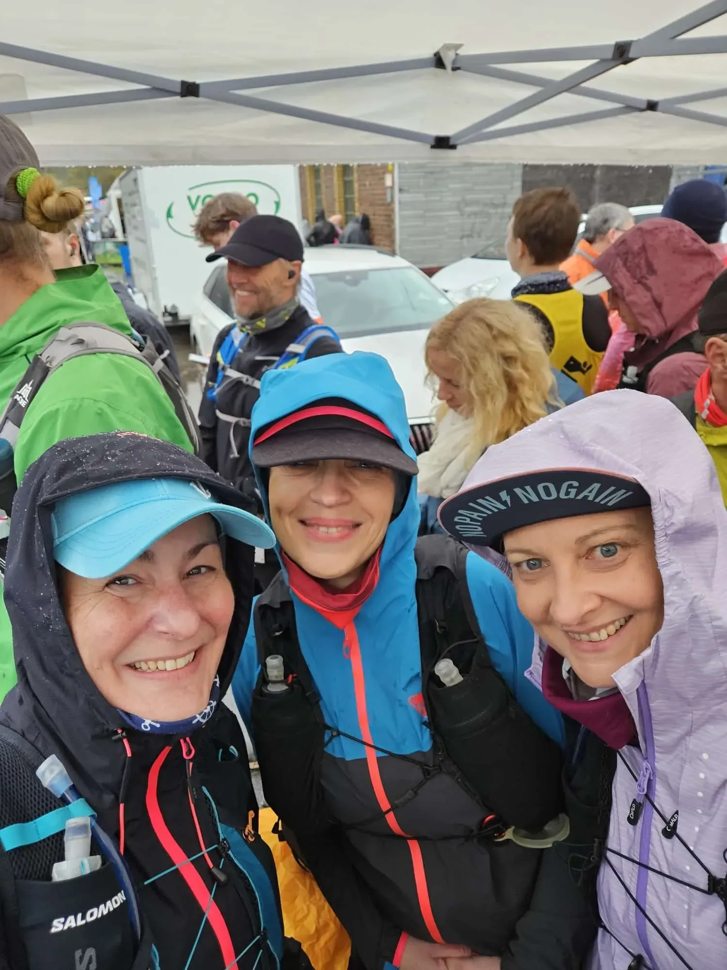 Drei Personen in Regenkleidung lächeln in die Kamera und warten bei einer Veranstaltung im Freien.