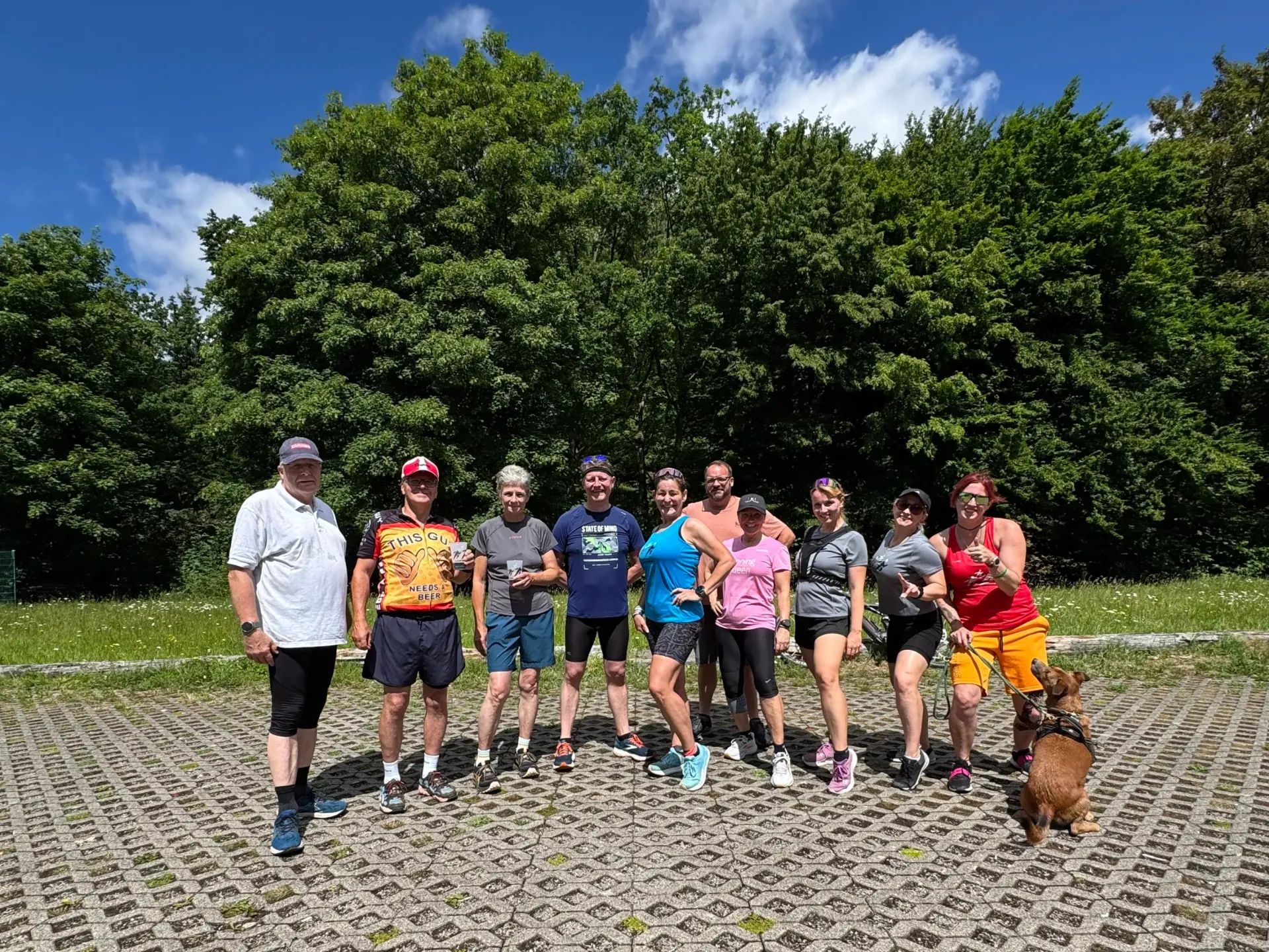 Eine Gruppe lächelnder Menschen steht im Freien. Hinter ihnen erstreckt sich eine Baumreihe unter blauem Himmel.