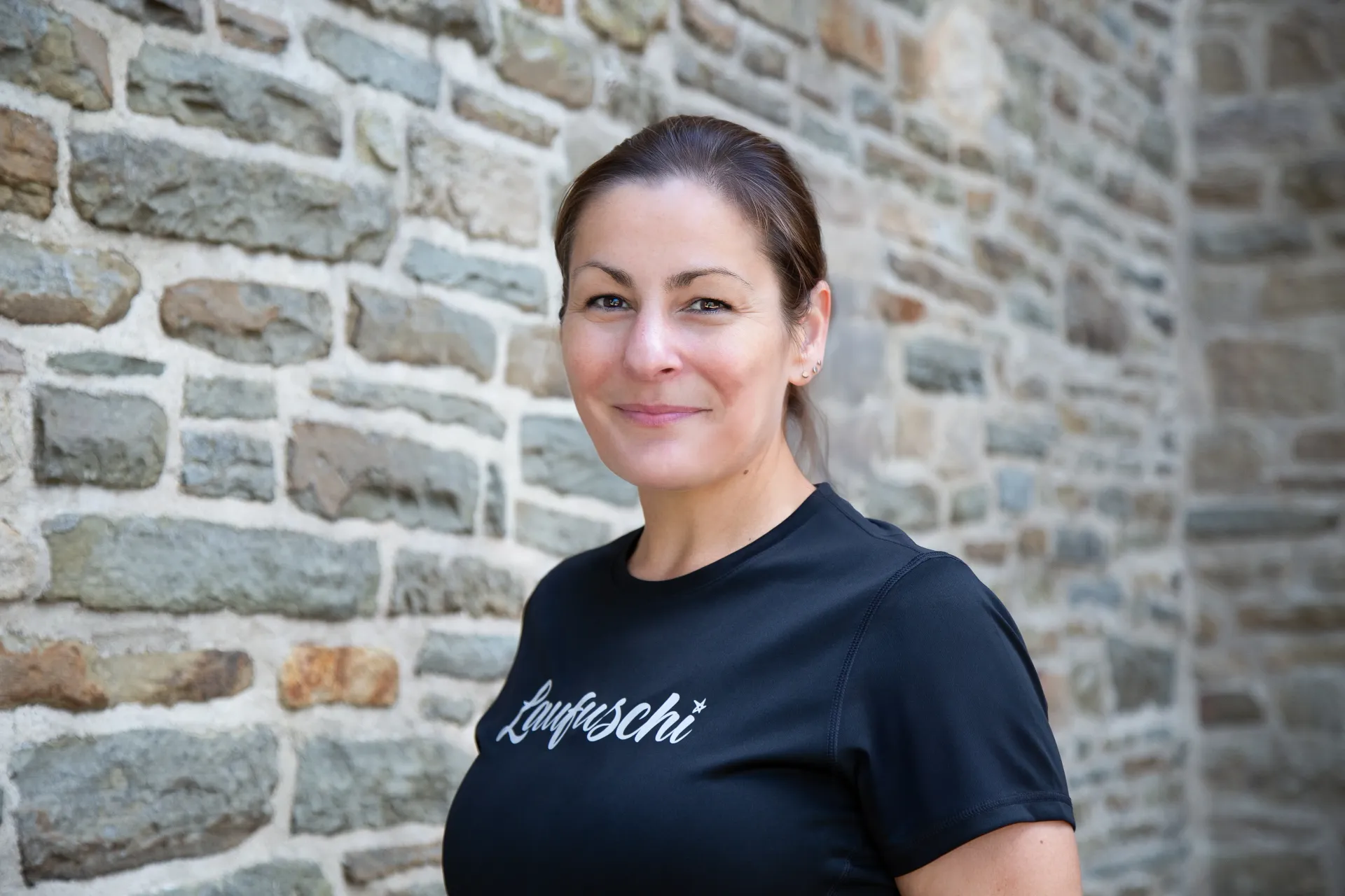 Eine Frau in schwarzem T-Shirt lächelt und lehnt an einer Steinmauer.