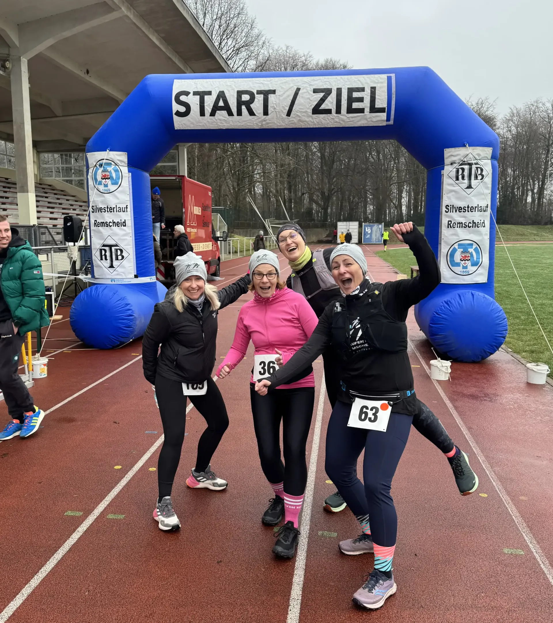 Vier Personen an der Ziellinie eines Rennens unter einem blauen Bogen mit der Aufschrift „START/ZIEL“. Sie lächeln und posieren.