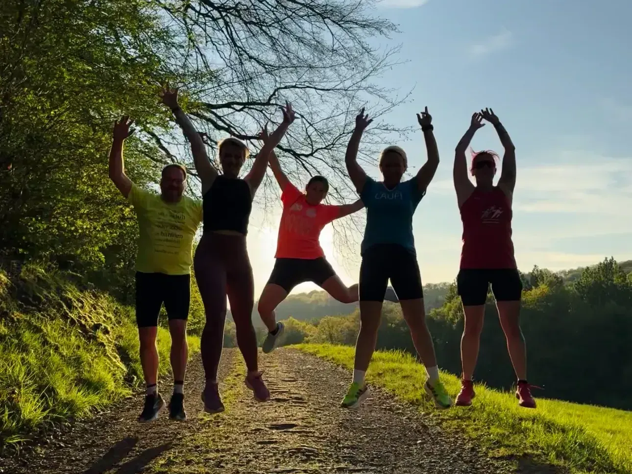 Fünf Personen springen mit erhobenen Armen auf einem sonnenbeschienenen Feldweg zwischen Bäumen.