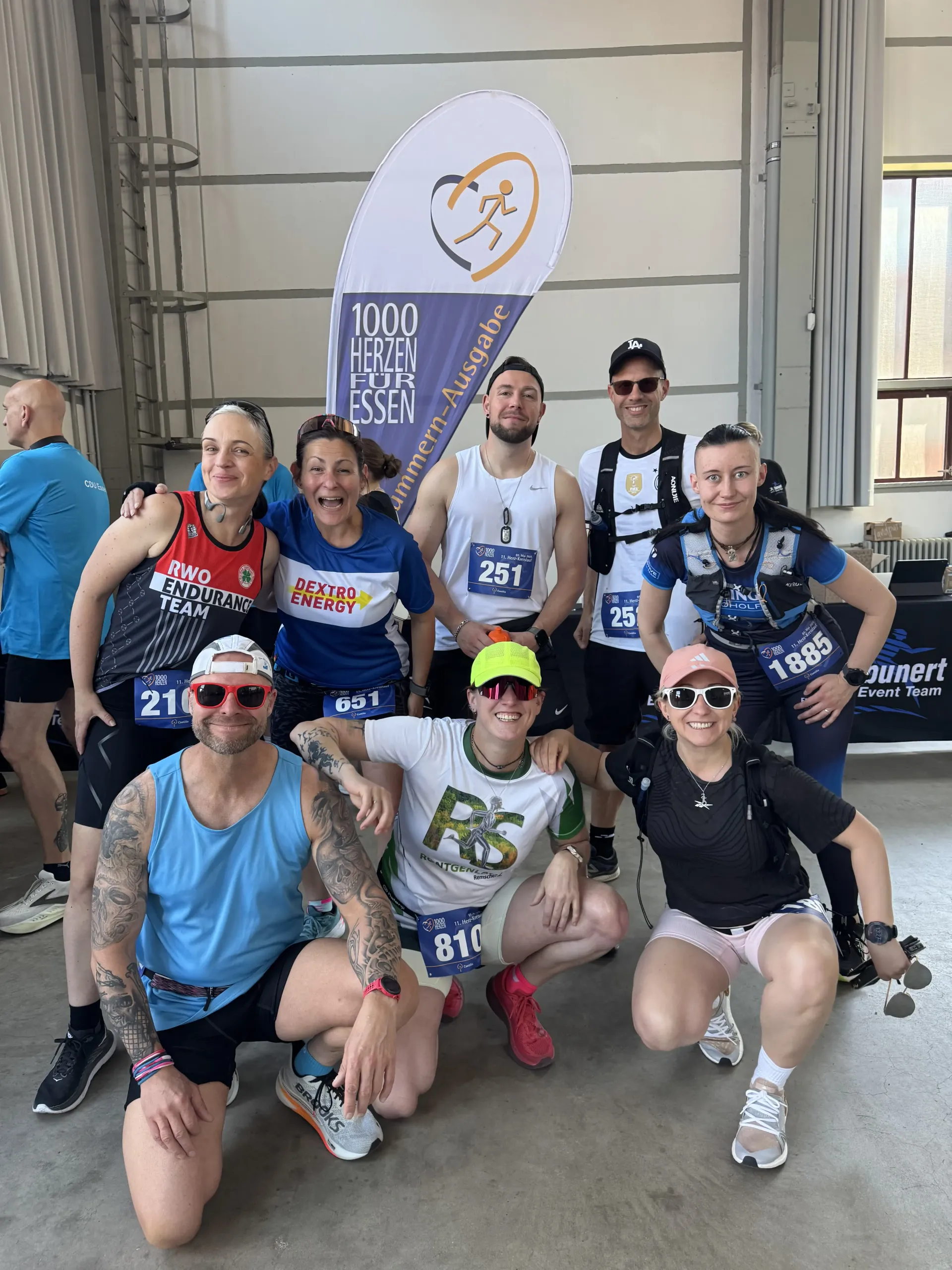 Eine Gruppe Läufer posiert mit einem Wettkampfbanner. Sie lächeln, tragen Sonnenbrillen, Sportkleidung und Startnummern. Wettkampf in einer Halle.