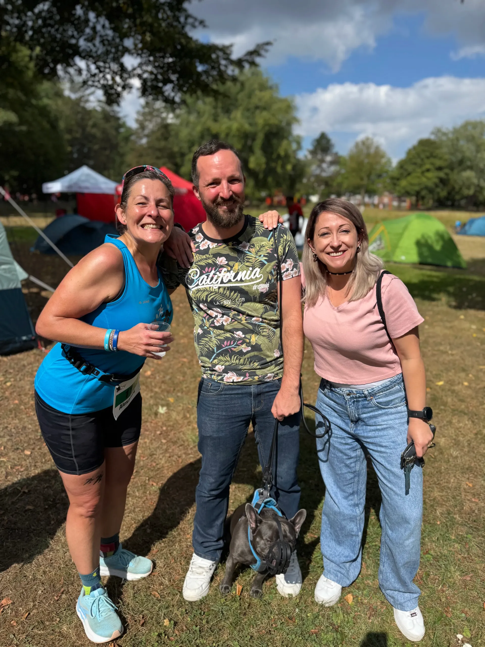 Three people smiling, posing with a dog in a grassy field with tents. One woman in athletic wear, two others in casual attire.