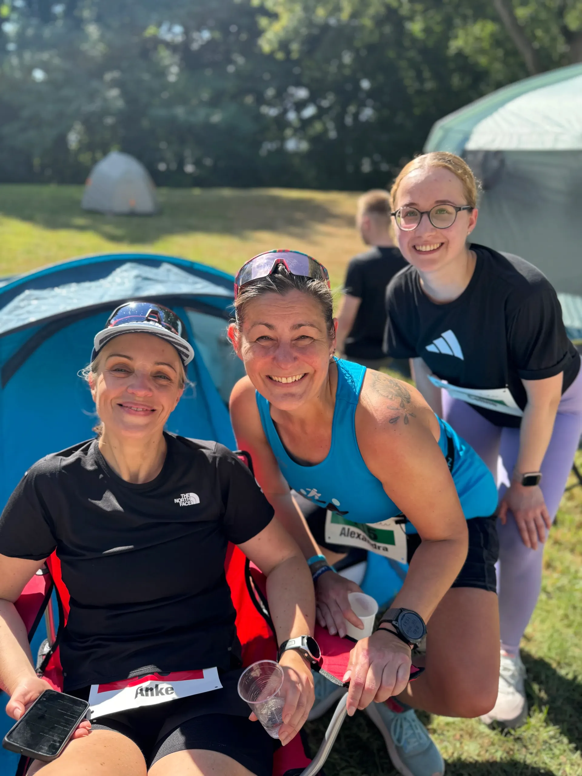 Three smiling women pose outdoors after a race, near a tent.
