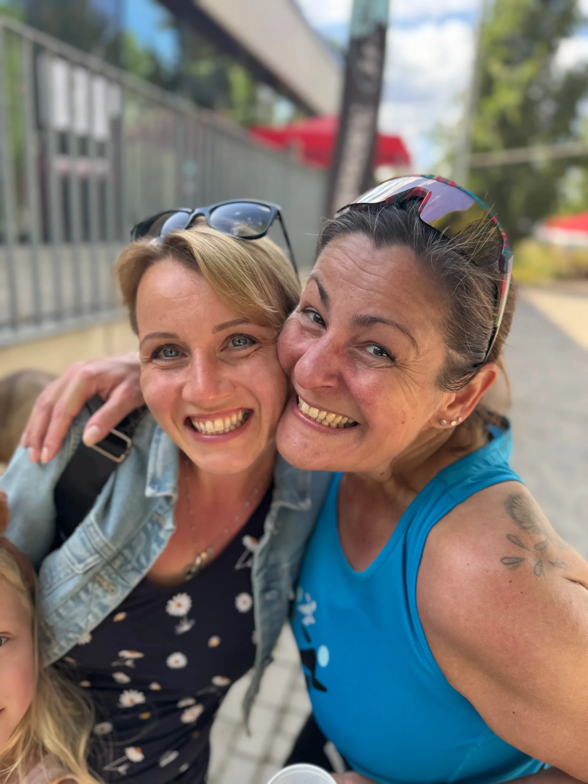 Two smiling women embracing, outdoors. One blonde, one brunette in a blue tank top.