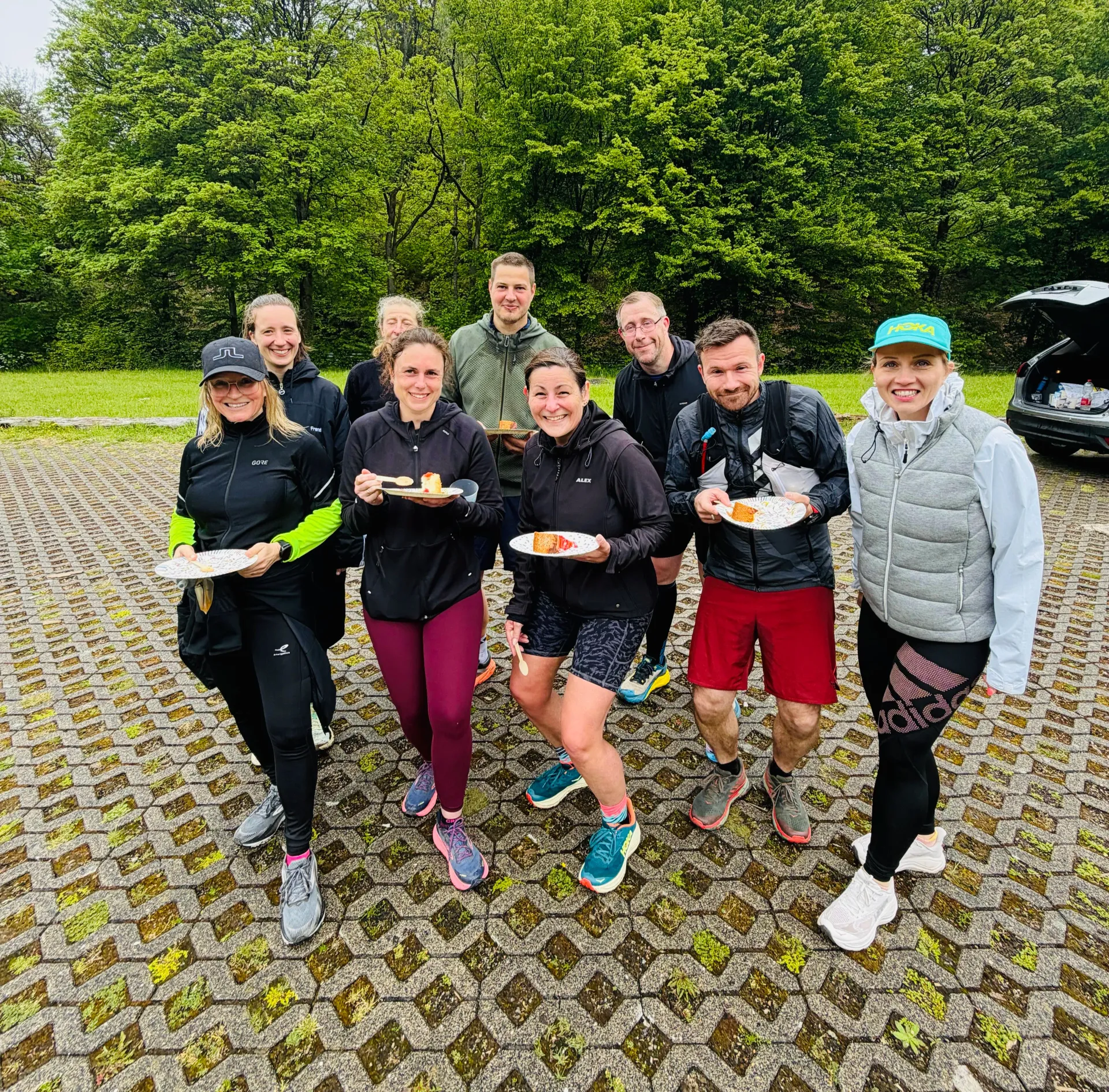 Eine Gruppe von Läufern lächelt im Freien auf einem nassen Steinweg, hält Getränke und Snacks in den Händen, im Hintergrund grüne Bäume.