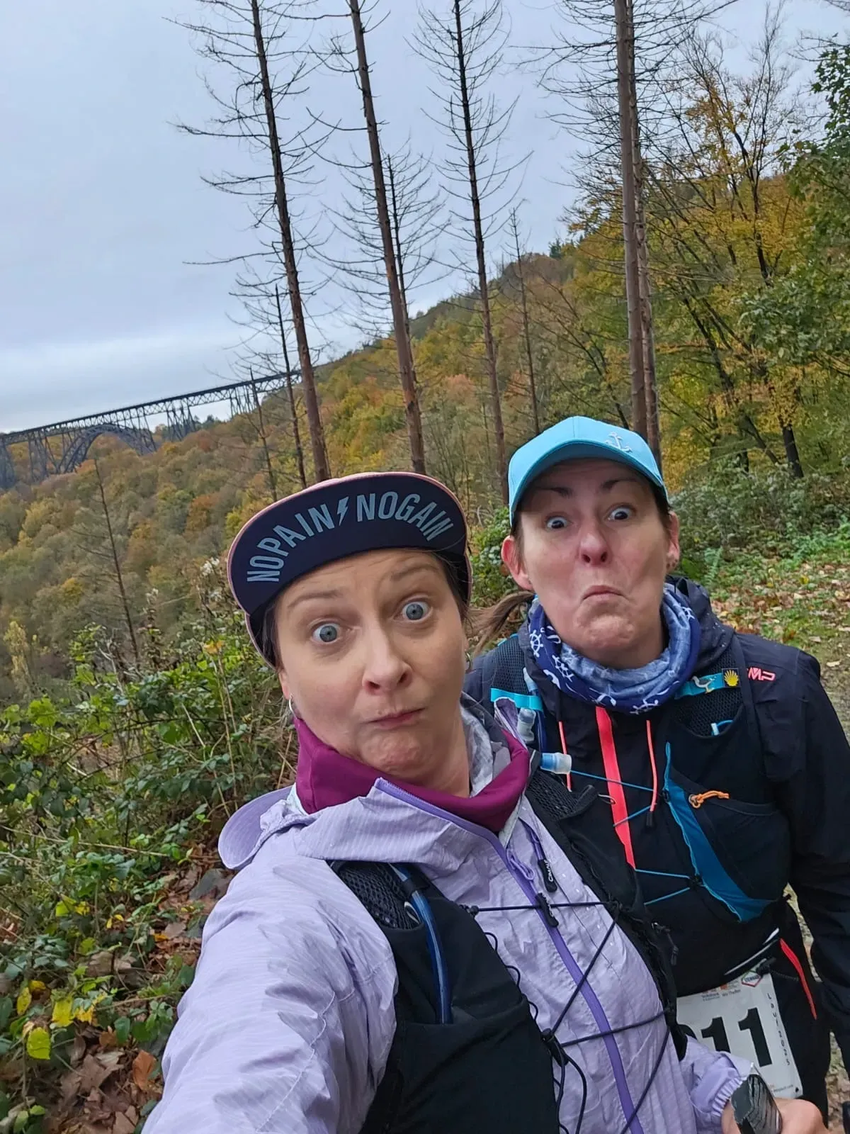 Zwei Frauen schneiden Grimassen und stehen auf einem Wanderweg. Herbstlaub und eine Brücke sind im Hintergrund zu sehen.