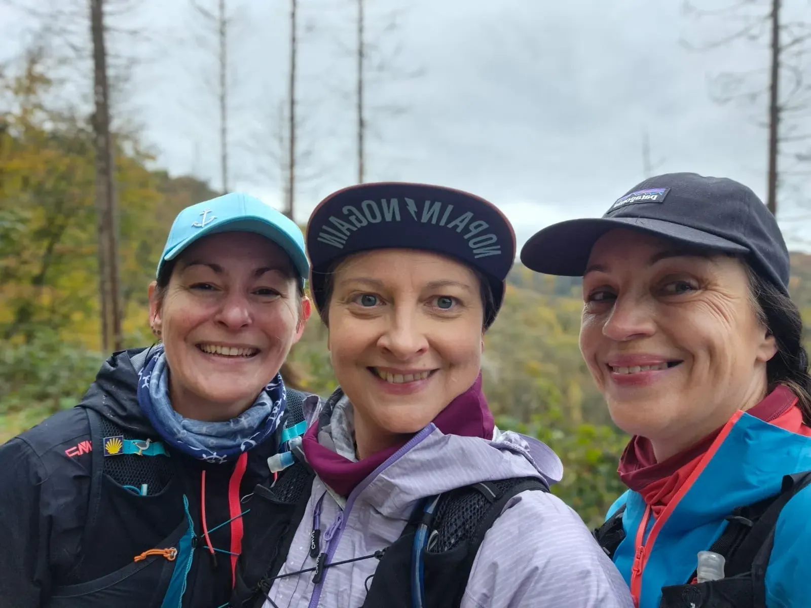 Drei Frauen lächeln und machen ein Selfie im Freien, umgeben von Bäumen.