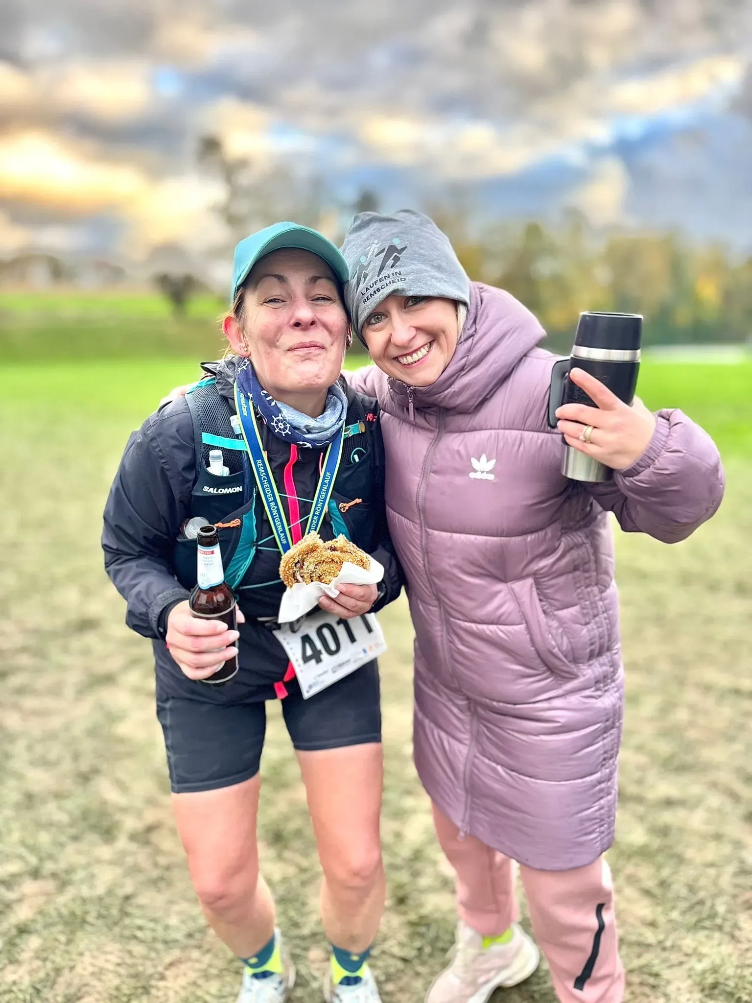 Zwei Frauen, die eine in Laufkleidung mit Medaille und Keks, die andere in einem Wintermantel, lächeln im Freien und halten eine Thermoskanne.