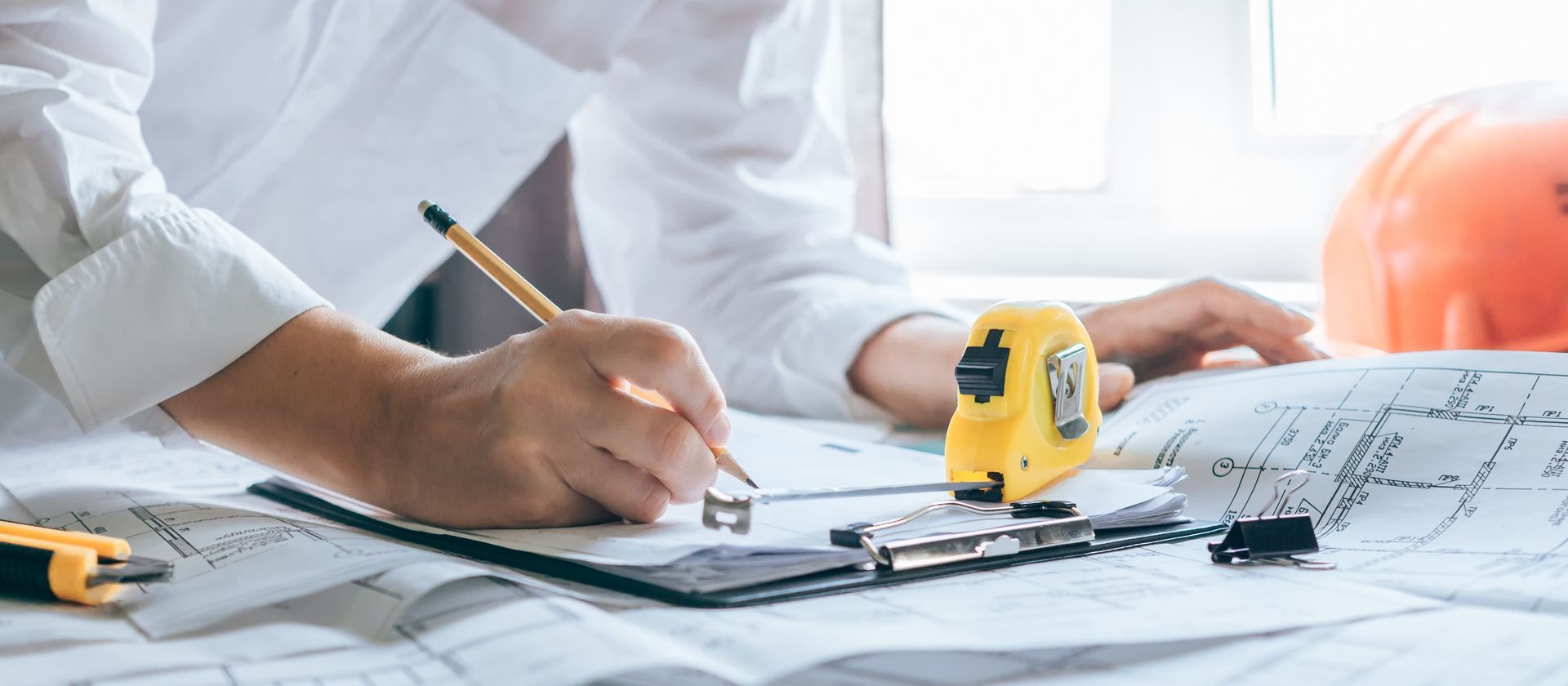Une personne dessine sur des plans avec un crayon, un mètre ruban et un casque de chantier, dans un environnement lumineux.