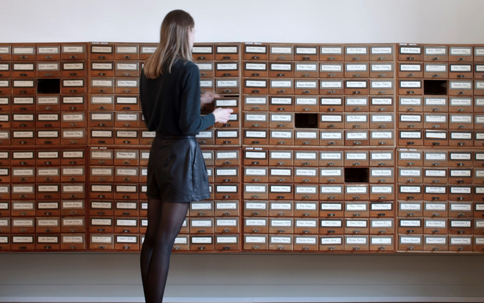 Bureau avec bibliothèque intégrée, bureau gris, deux bustes, porte. Chaise marron, parquet, couleurs neutres.