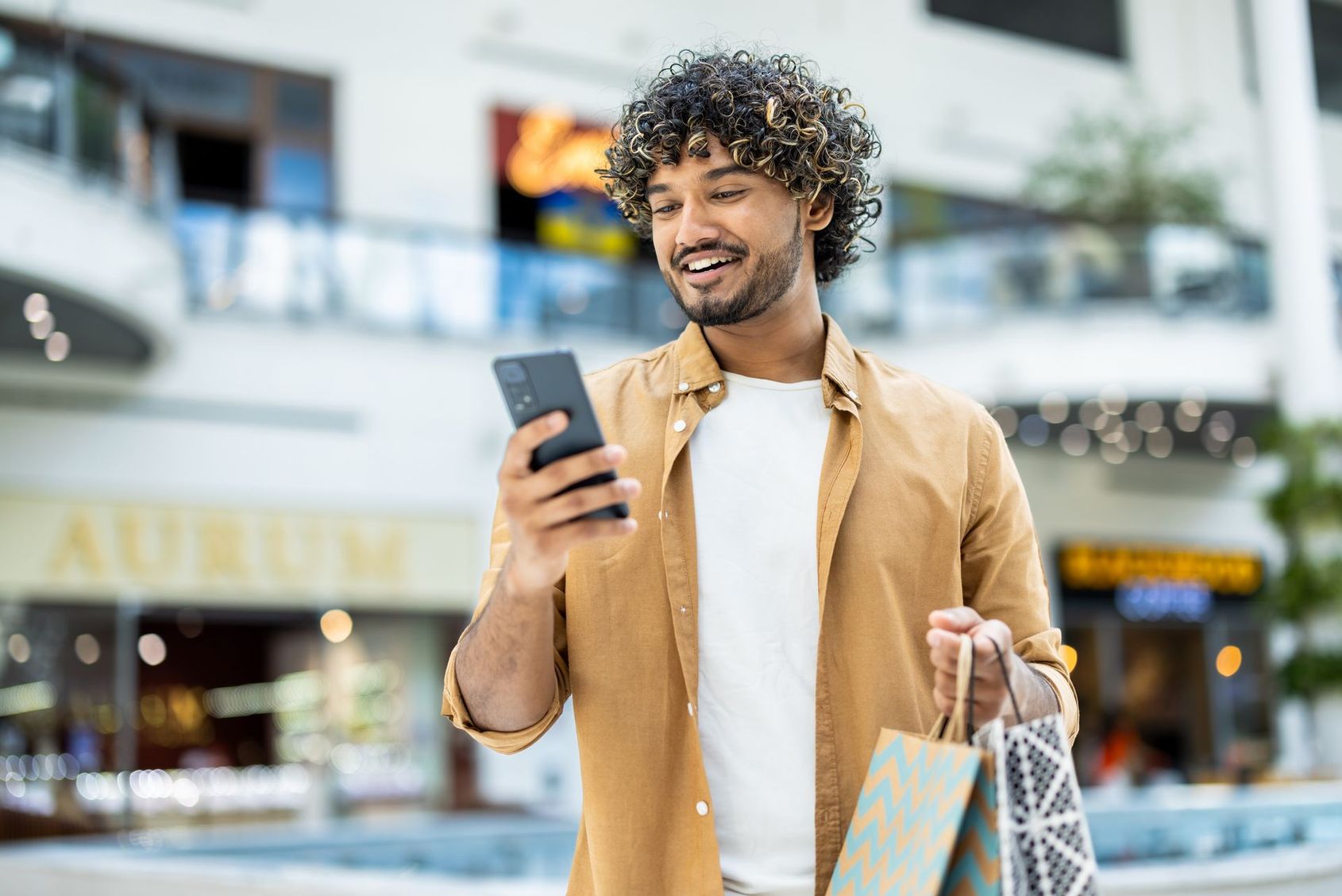 Un homme souriant avec des sacs de shopping regarde son téléphone dans une galerie marchande
