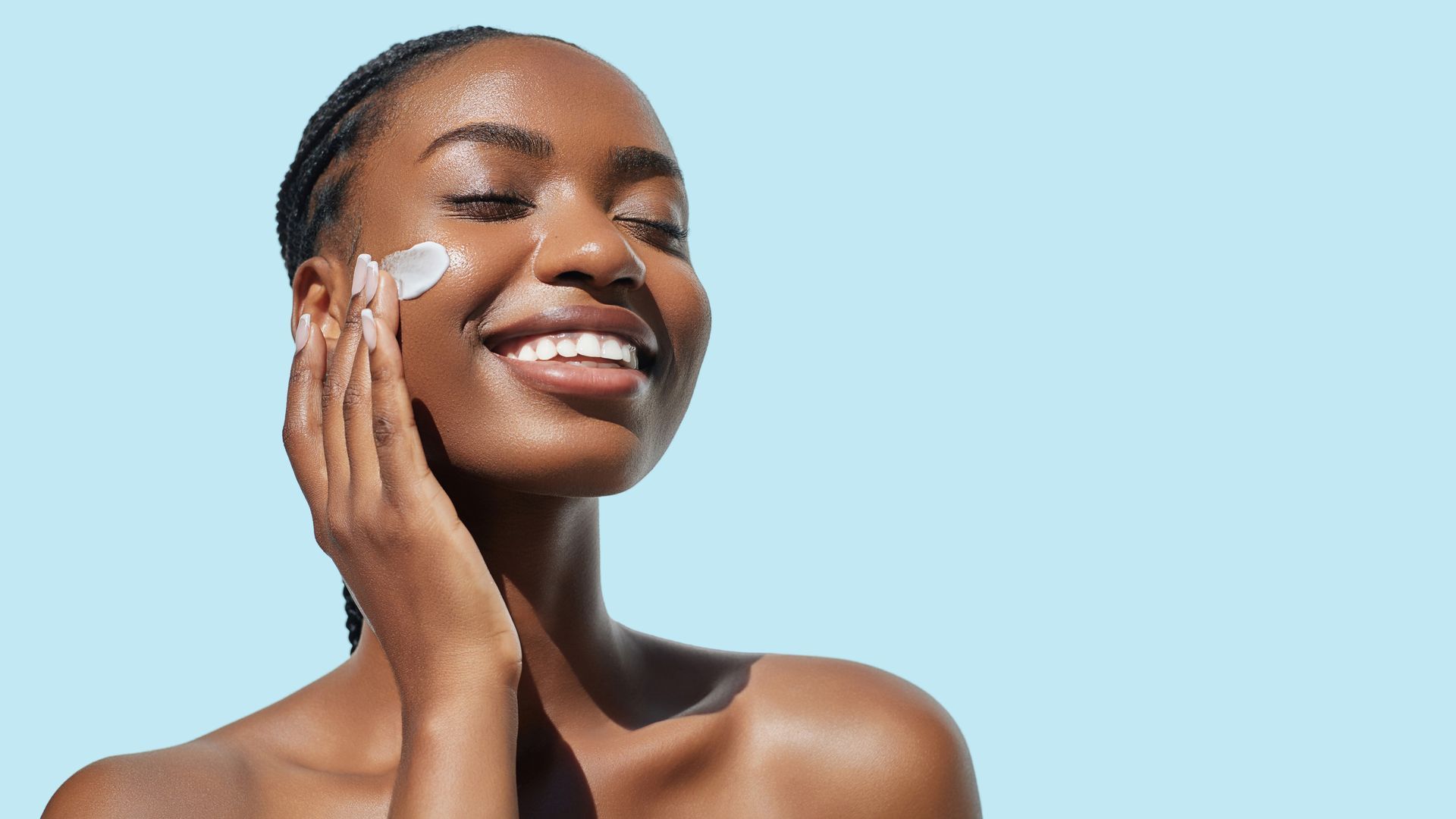 Personne souriante appliquant de la crème sur la joue, sur fond bleu clair