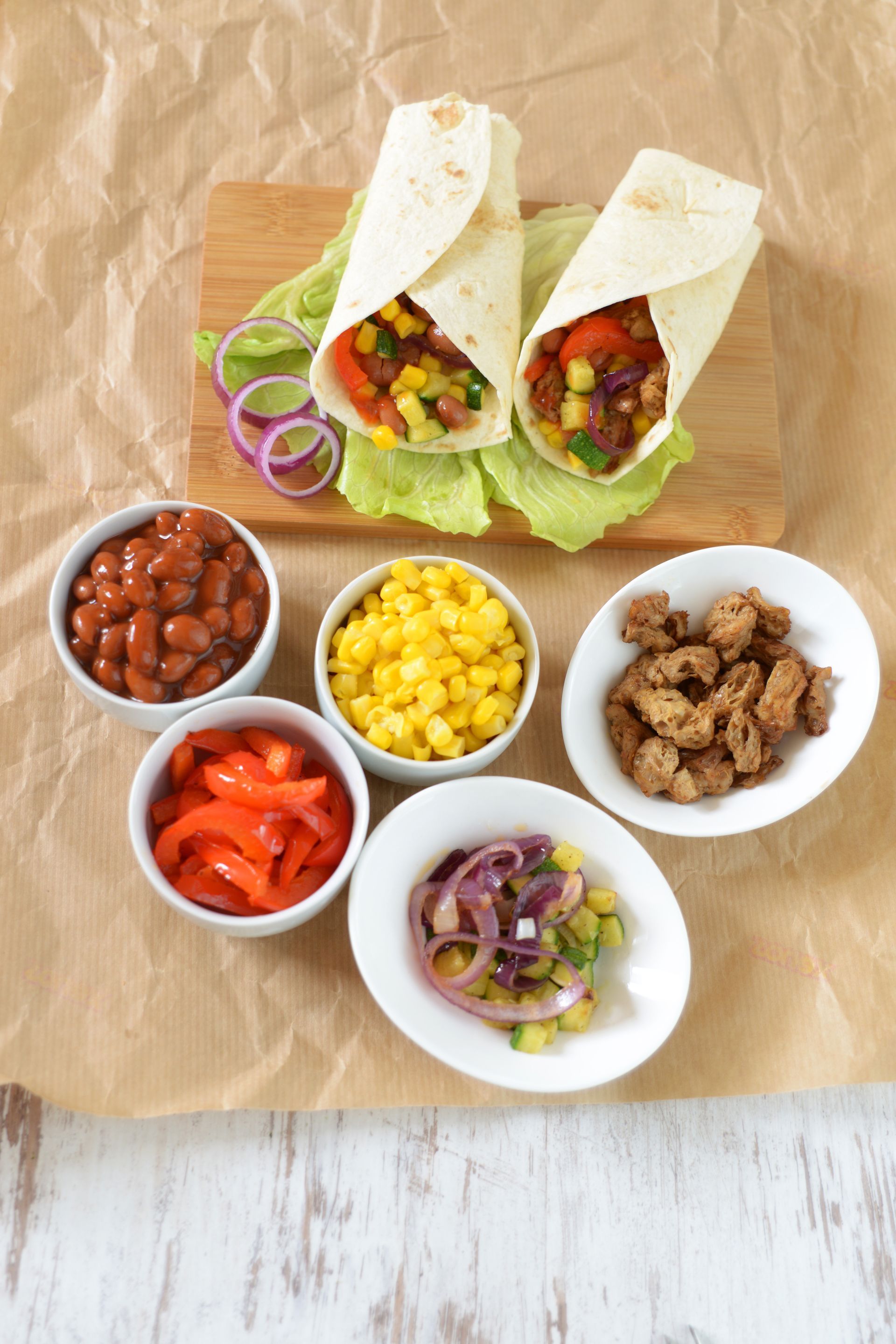 Garniture pour tacos avec deux tacos emballés et des bols de haricots, de maïs, de tomates, d’oignons et de viande sur du papier brun.