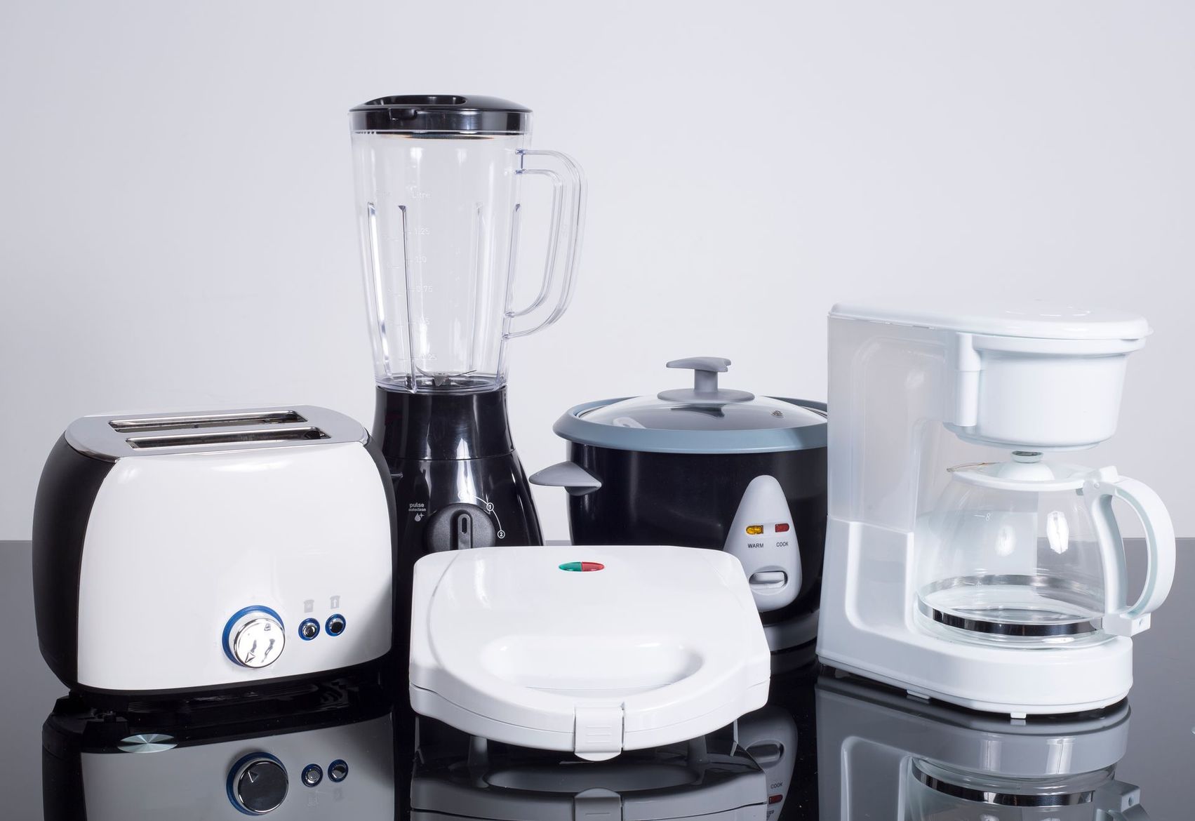 Divers appareils de cuisine blancs disposés sur une surface noire brillante, dont un mixeur, un grille-pain, une cafetière et un appareil à croque-monsieur.
