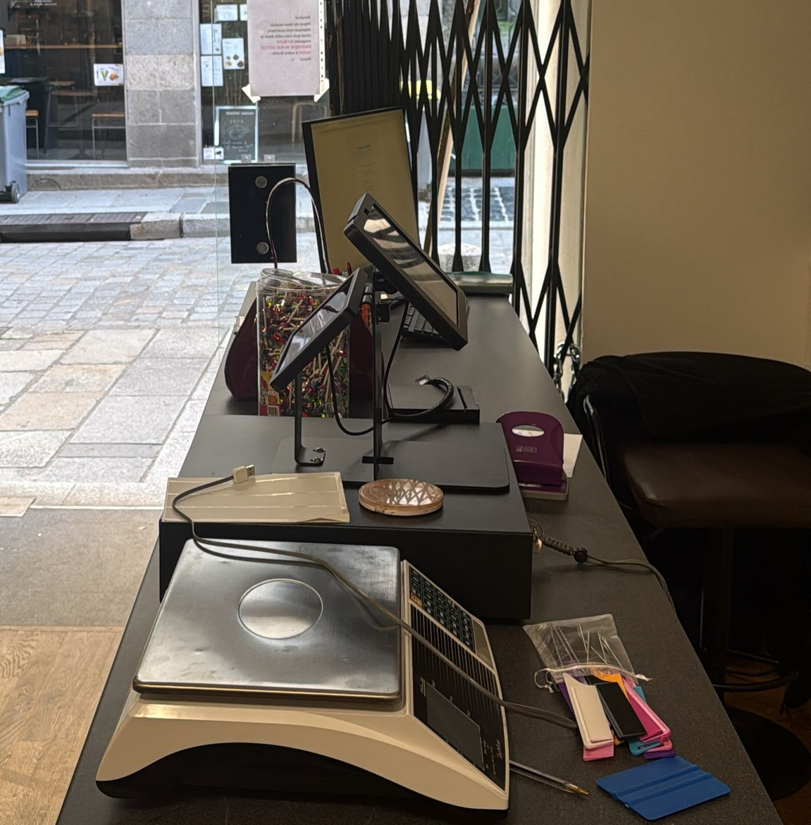 Petit comptoir de café avec une caisse enregistreuse, donnant sur une rue pavée de pierres à l’extérieur.