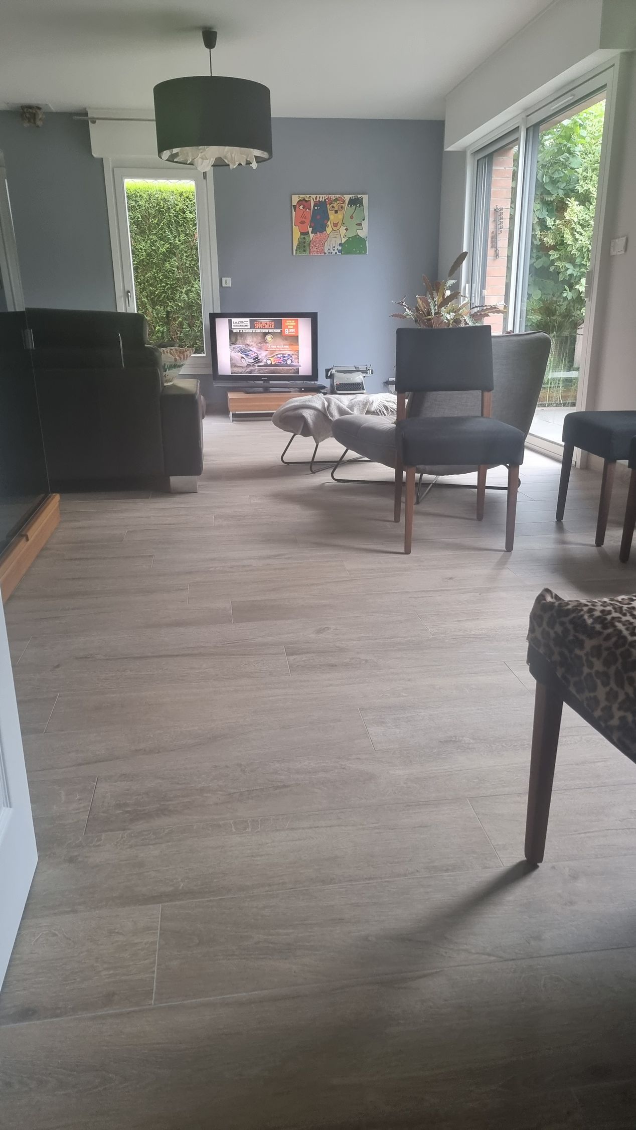 Salon moderne avec murs gris, grandes fenêtres, fauteuils, télévision et parquet.