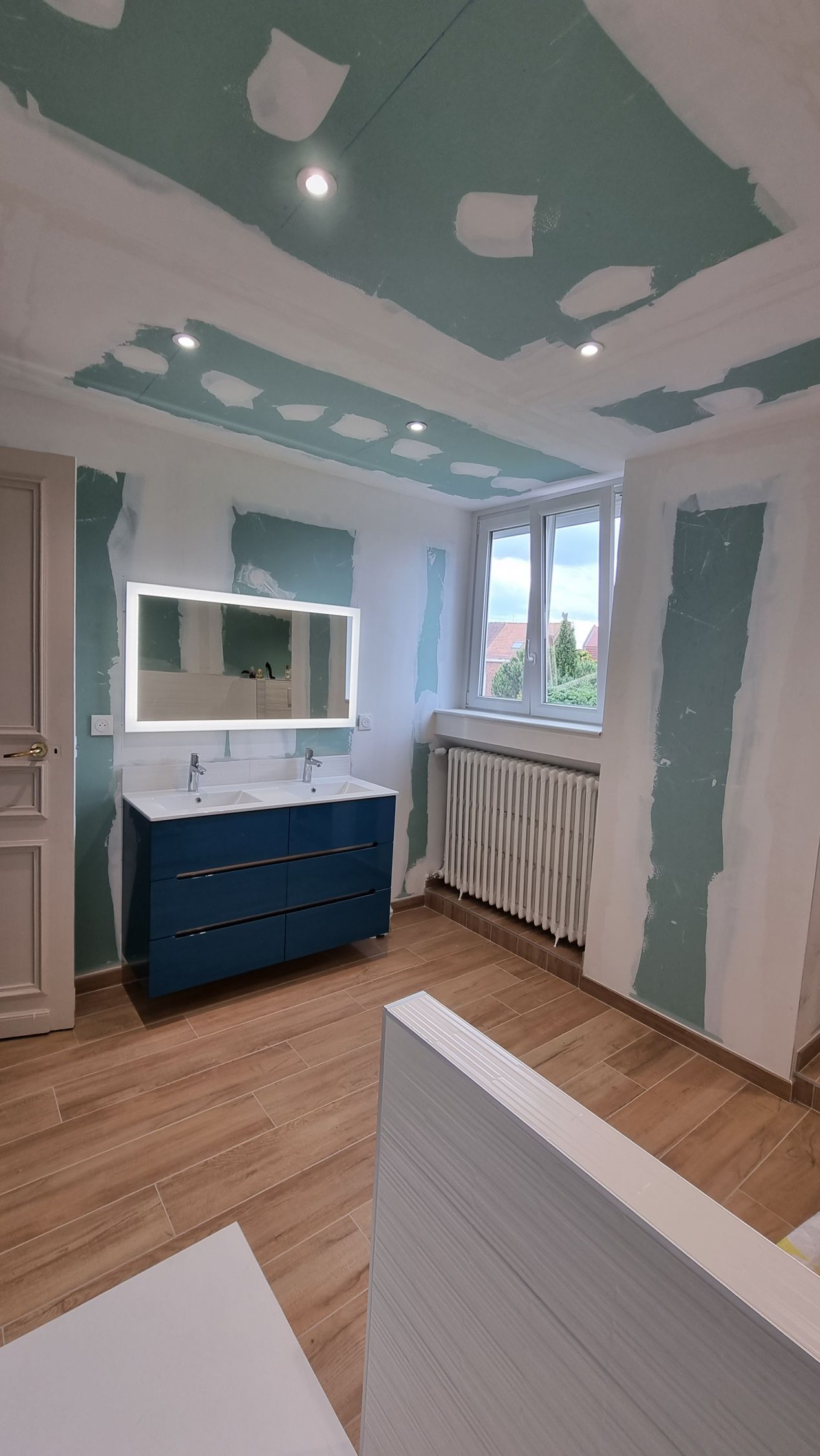 Chambre partiellement rénovée avec meuble-lavabo bleu, touches de bleu sarcelle au plafond, fenêtre et radiateur en construction