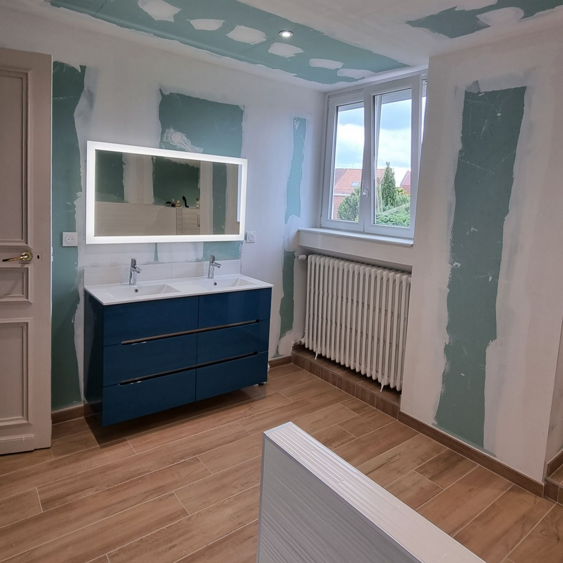 Petite salle de bains rénovée avec meuble-lavabo bleu, miroir, fenêtre, radiateur et parquet clair.