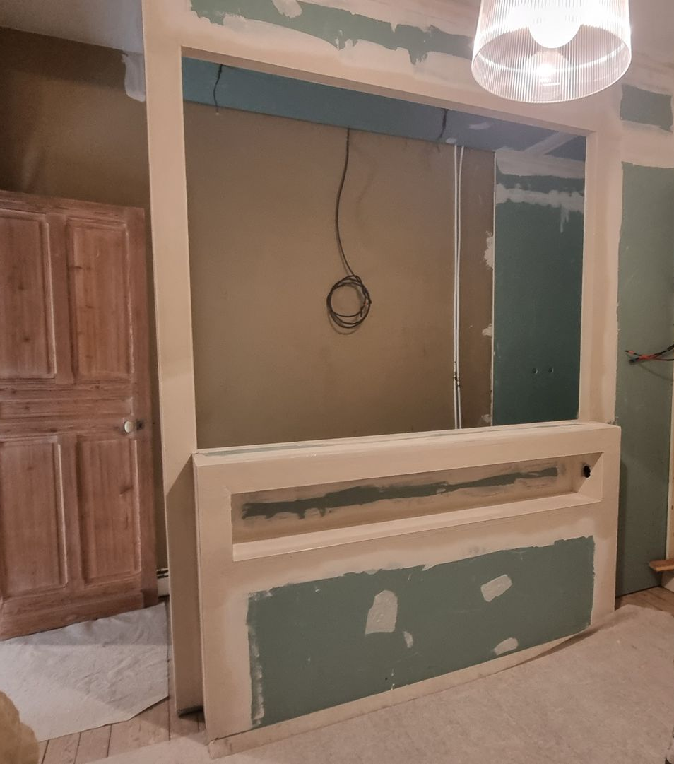 Ouverture murale inachevée avec des plaques de plâtre réparées, des câbles apparents et un luminaire suspendu dans une pièce en rénovation