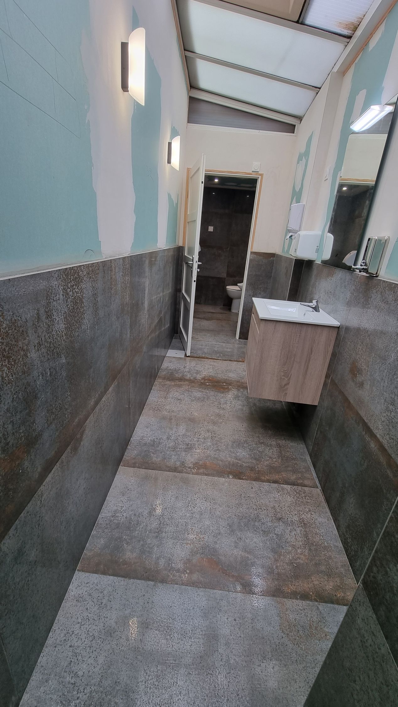 Couloir intérieur étroit avec murs carrelés, puits de lumière et un petit meuble-lavabo sur la droite.