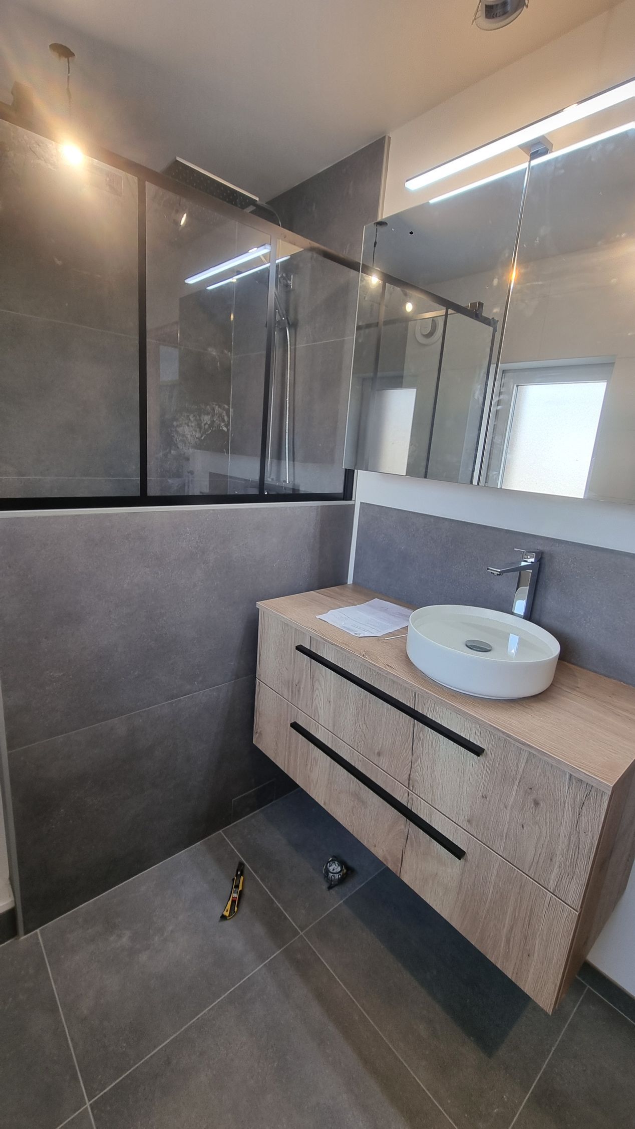 Salle de bains moderne grise avec douche en verre, miroir mural et meuble-lavabo en bois avec lavabo rond.