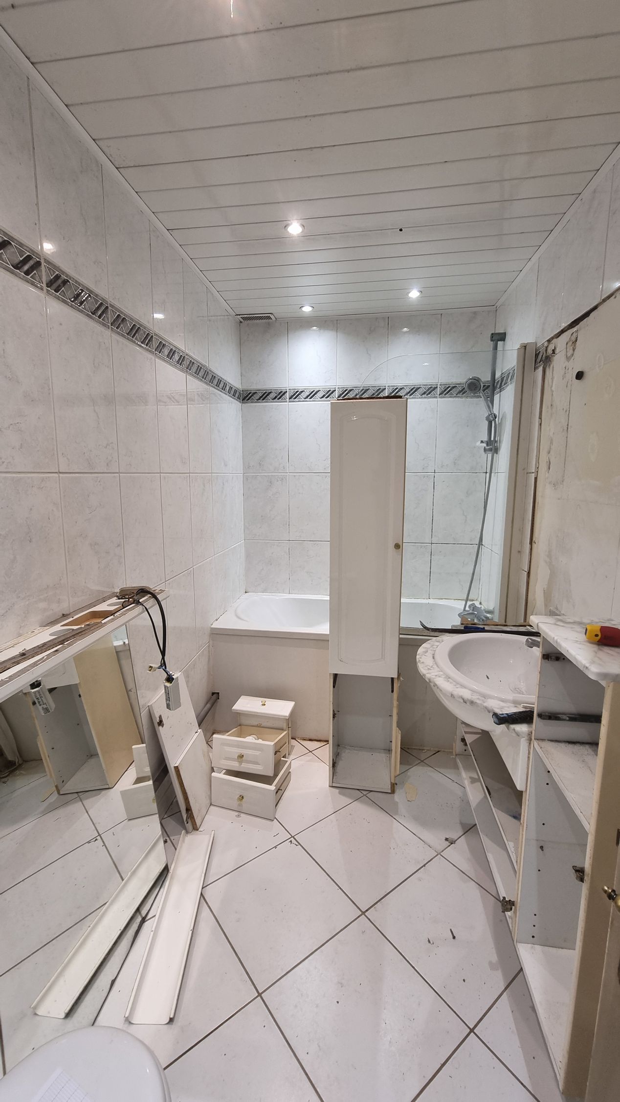 Petite salle de bains carrelée avec douche, lavabo, toilettes et miroir, éclairée par un plafonnier lumineux.