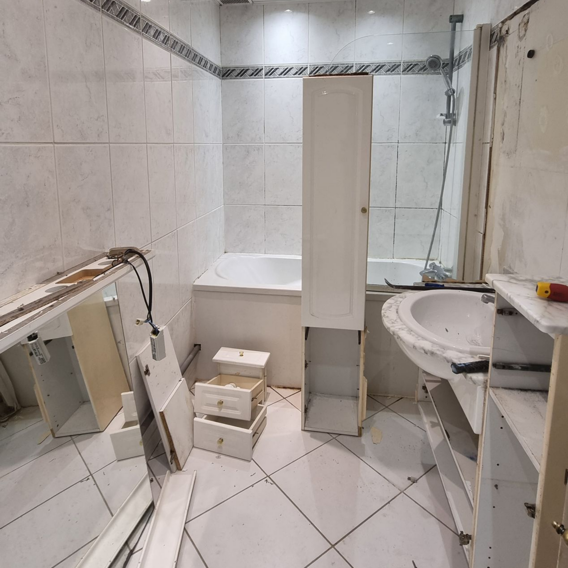 Petite salle de bains encombrée avec des carreaux blancs, une baignoire, un lavabo et des morceaux de meubles cassés au sol.