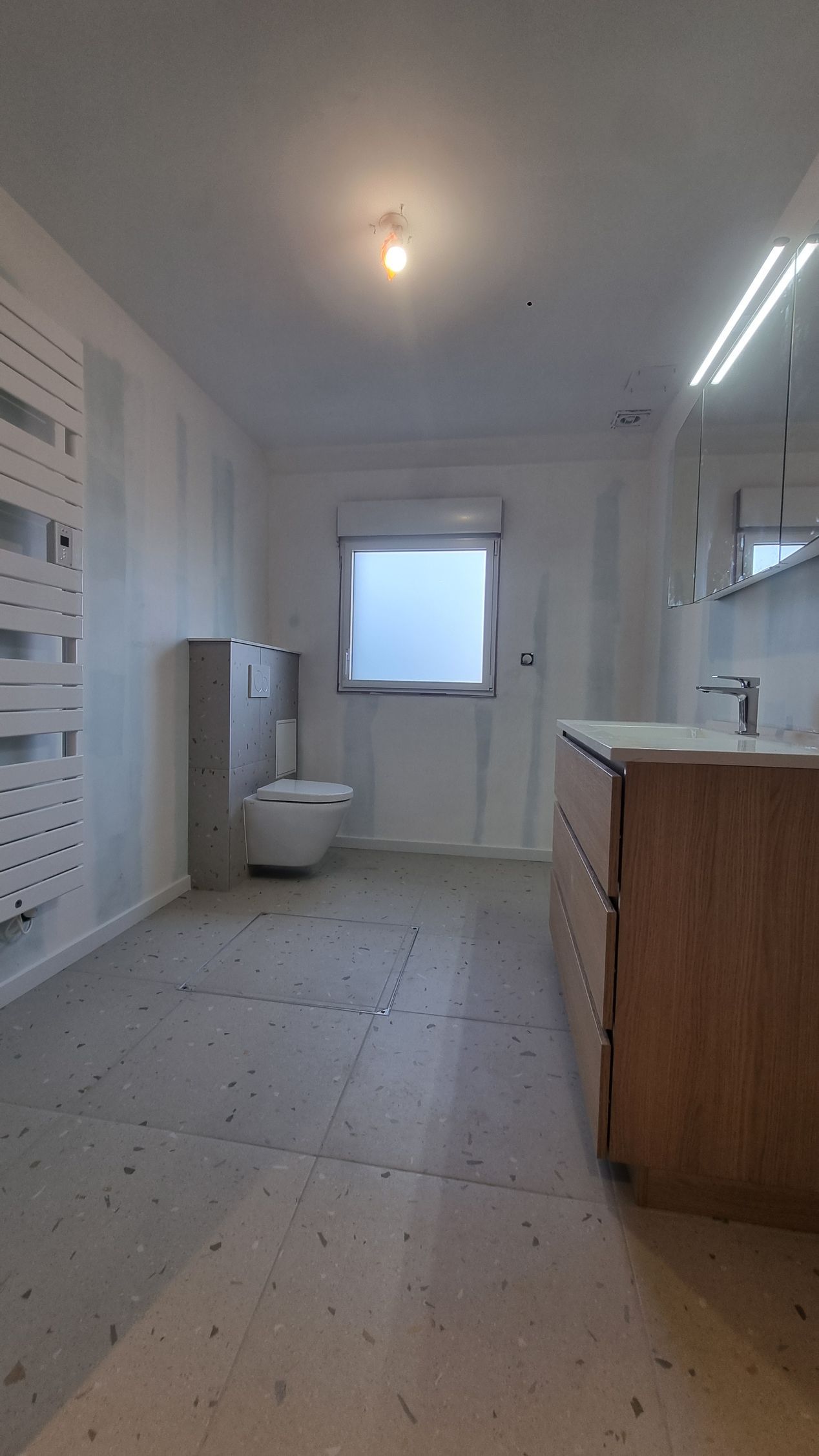 Cuisine blanche lumineuse avec sol carrelé, toilettes à gauche, fenêtre en face et îlot central en bois à droite.