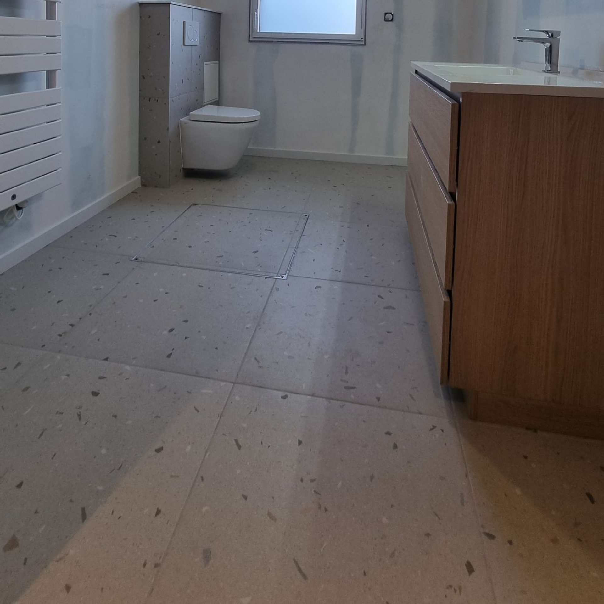Salle de bains moderne avec toilettes blanches, fenêtre, lavabo et meuble-lavabo en bois sur un sol carrelé moucheté
