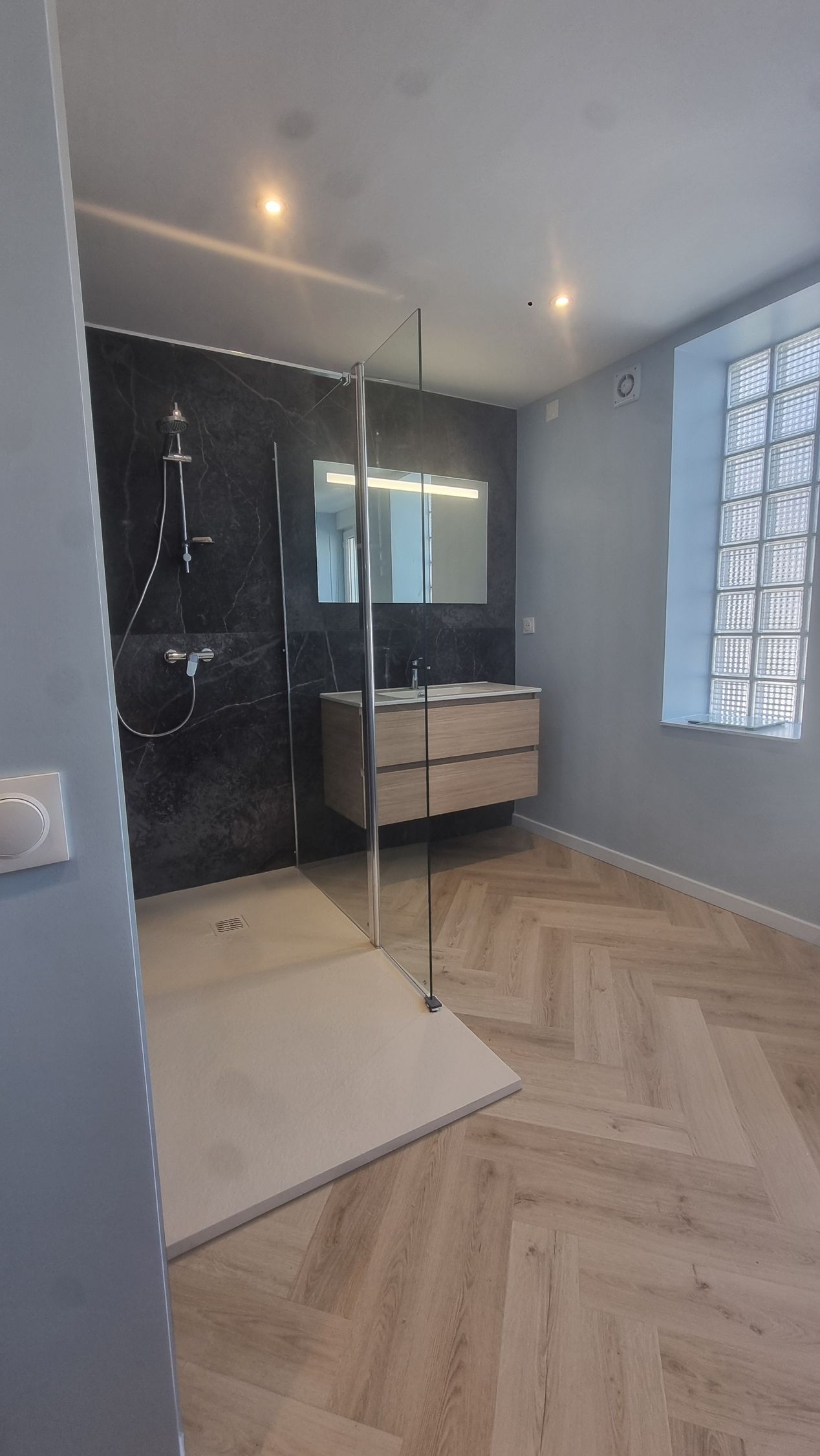 Salle de bains moderne avec douche noire, paroi en verre, meuble-lavabo, sol à chevrons et mur en pavés de verre