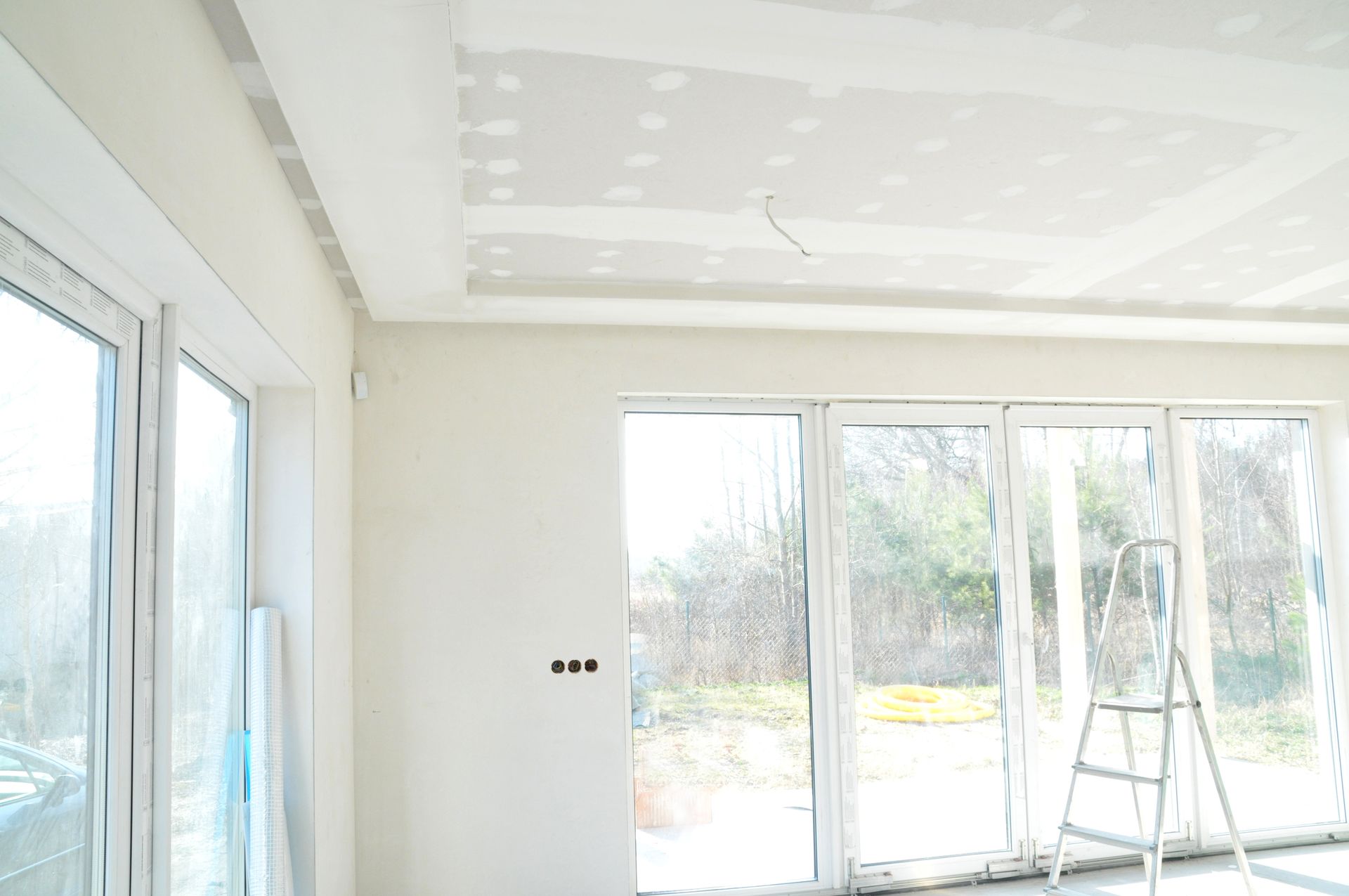 Pièce lumineuse non finie avec de grandes fenêtres, des murs blancs et une échelle près des portes vitrées
