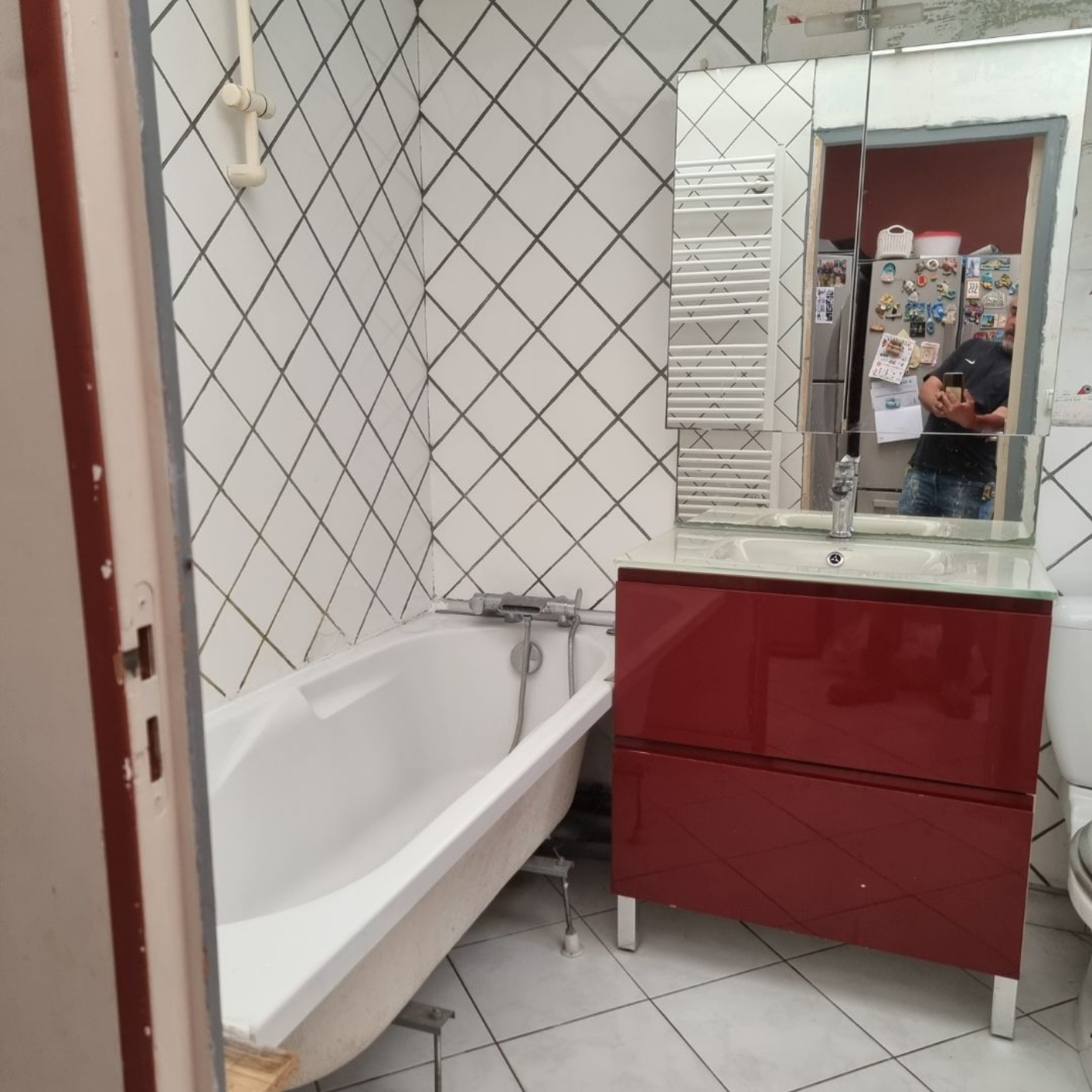 Salle de bains avec murs carrelés blancs, baignoire, meuble-lavabo rouge et miroir reflétant une personne prenant la photo
