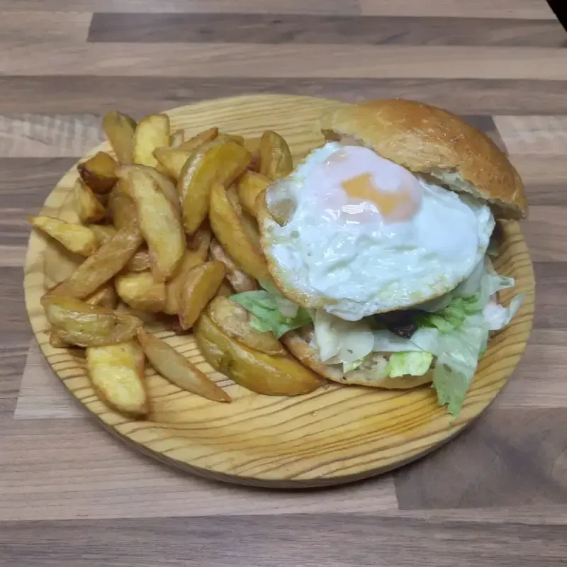 Hamburguesa con huevo frito y lechuga en una tabla de madera, servida con patatas fritas gruesas.