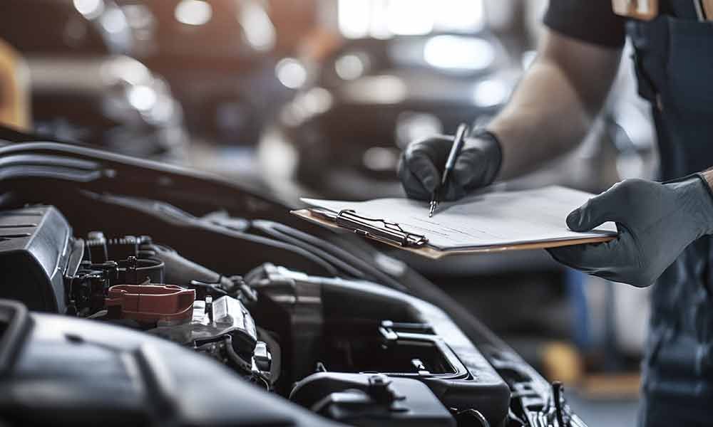 Ein Mechaniker inspiziert einen Automotor, schreibt dabei auf einem Klemmbrett in einer Werkstatt und trägt Handschuhe.