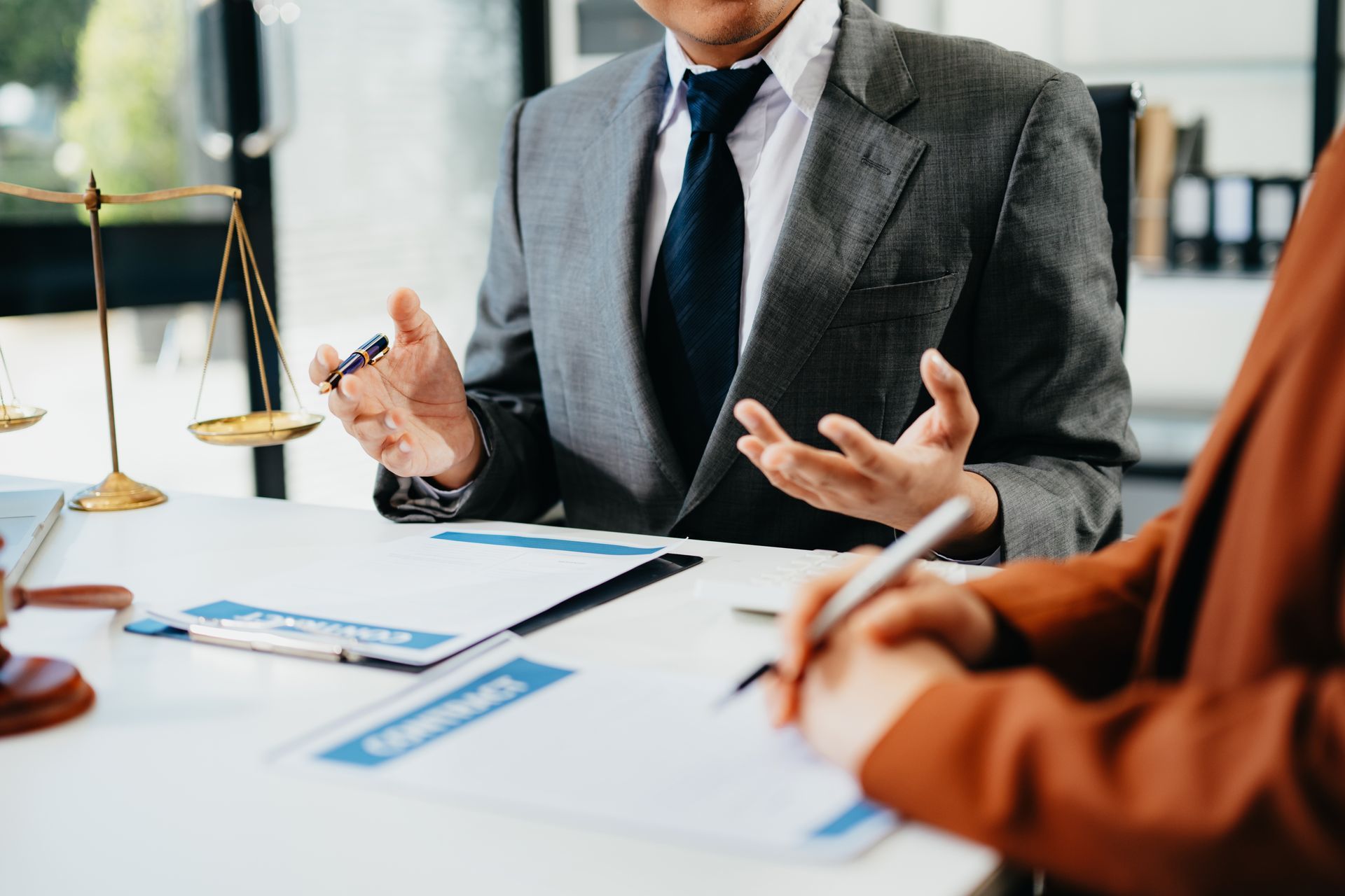 Un avocat en costume gesticule tout en discutant d'un contrat avec un client, assis à un bureau où se trouve une balance juridique.