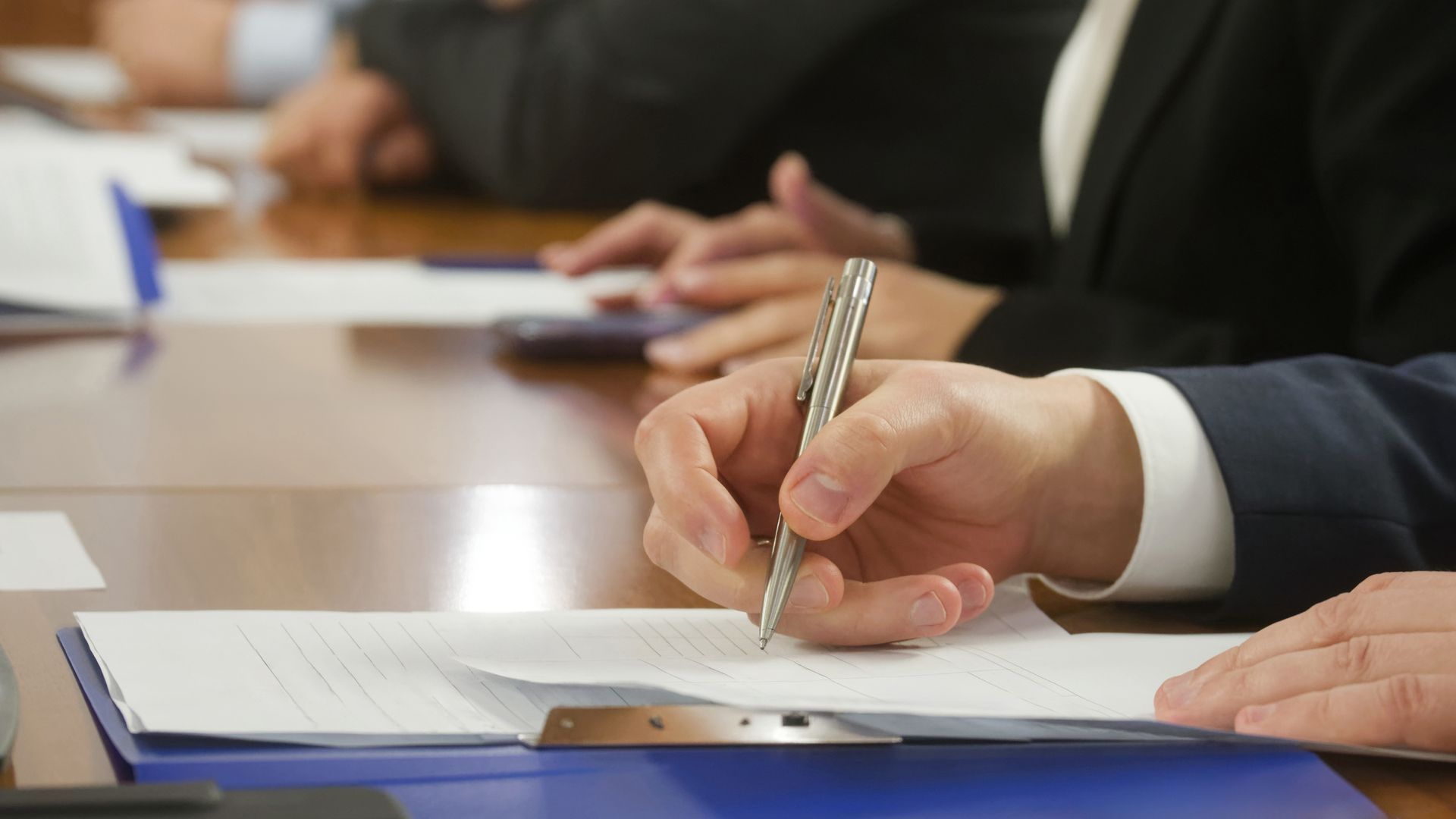 Gros plan sur la main d'une personne signant un document avec un stylo argenté sur un bureau lors d'une réunion professionnelle.