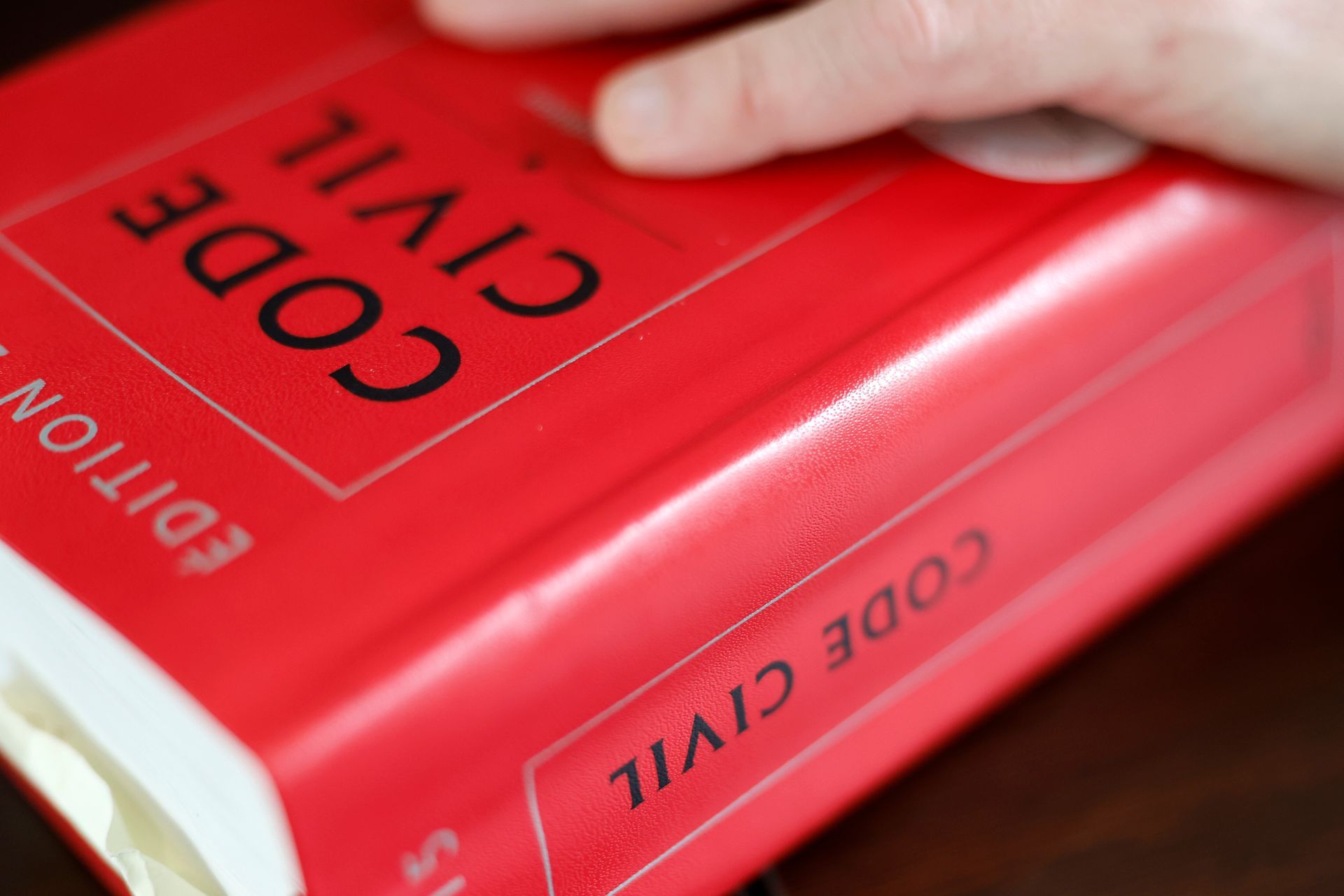 Une main repose sur un livre rouge vif intitulé CODE CIVIL, un code juridique français, posé sur une surface sombre.