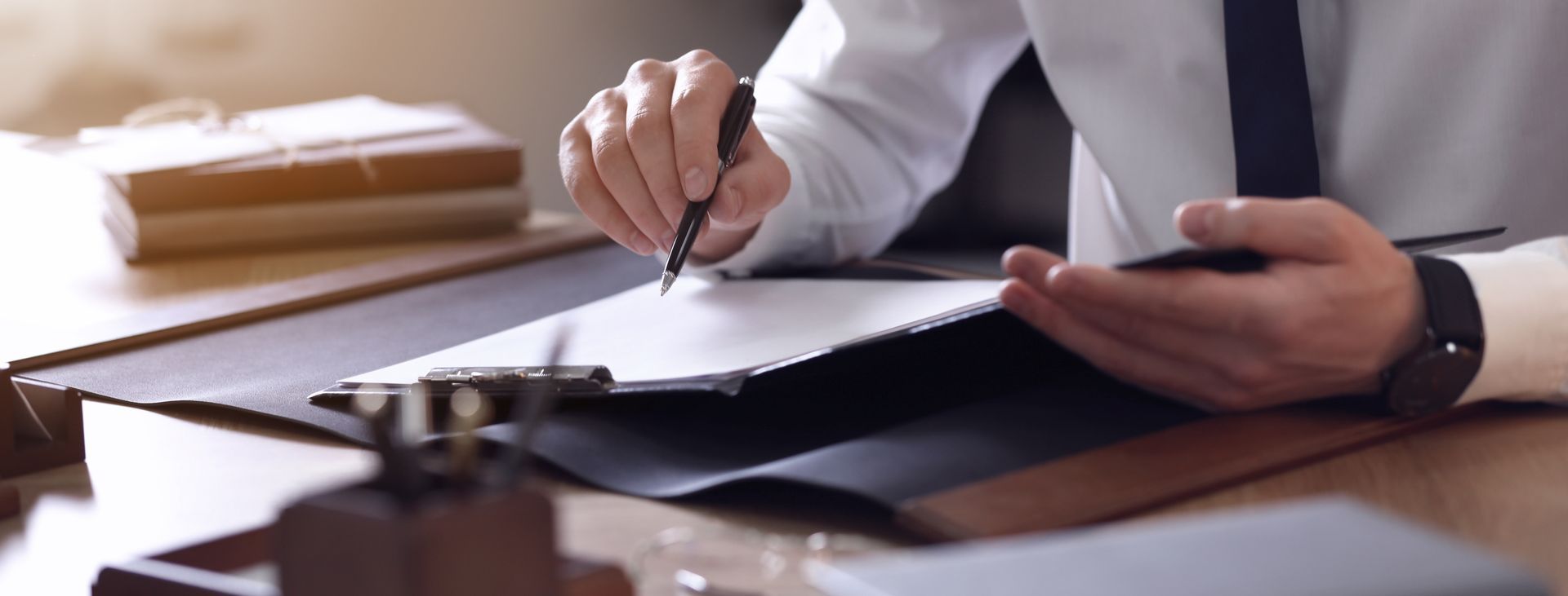 Une personne en chemise blanche et cravate, assise à un bureau, tenant un stylo au-dessus d'une feuille de papier posée sur un bloc-notes.